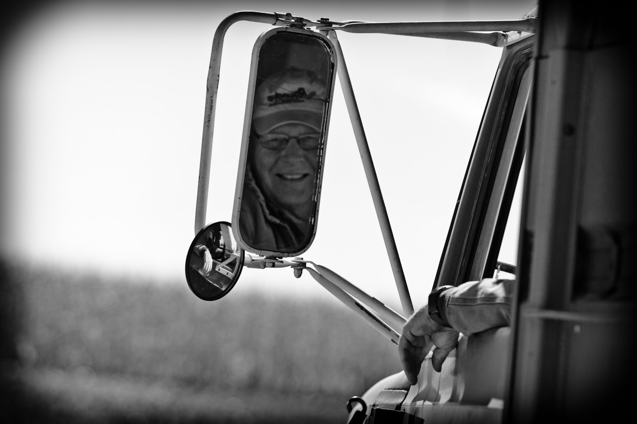 A sidel mirror of a truck shows the reflection of a smiling elderly man wearing glasses.