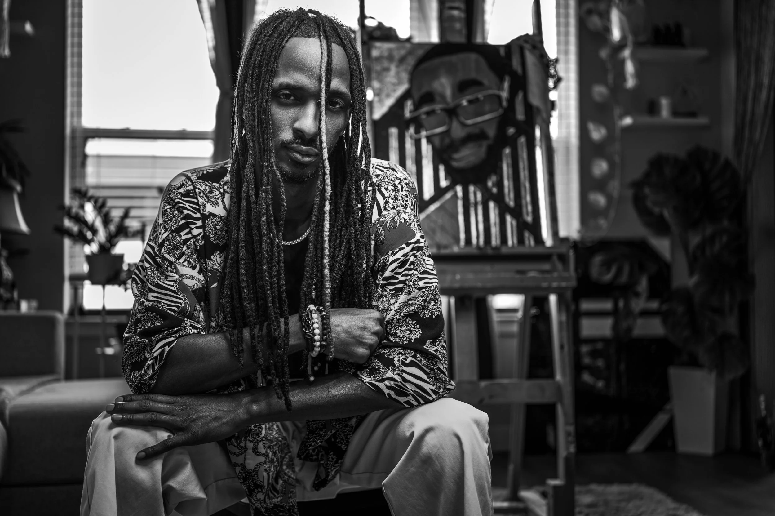 A young man with long dreadlocks and artistic clothing sitting with arms crossed in a creative studio or art space, with art supplies and masks visible in the background.