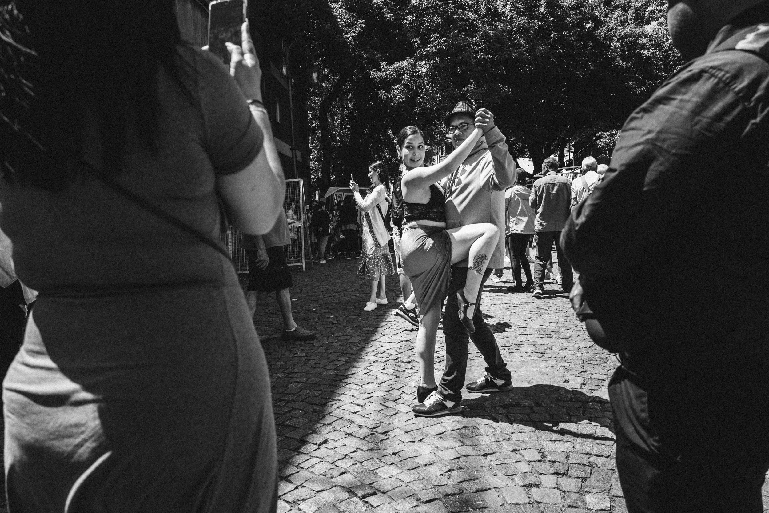 A black-and-white photo of a couple dancing Tango outdoors in Buenos Aires during a lively event, surrounded by people on a cobblestone street with trees in the background.