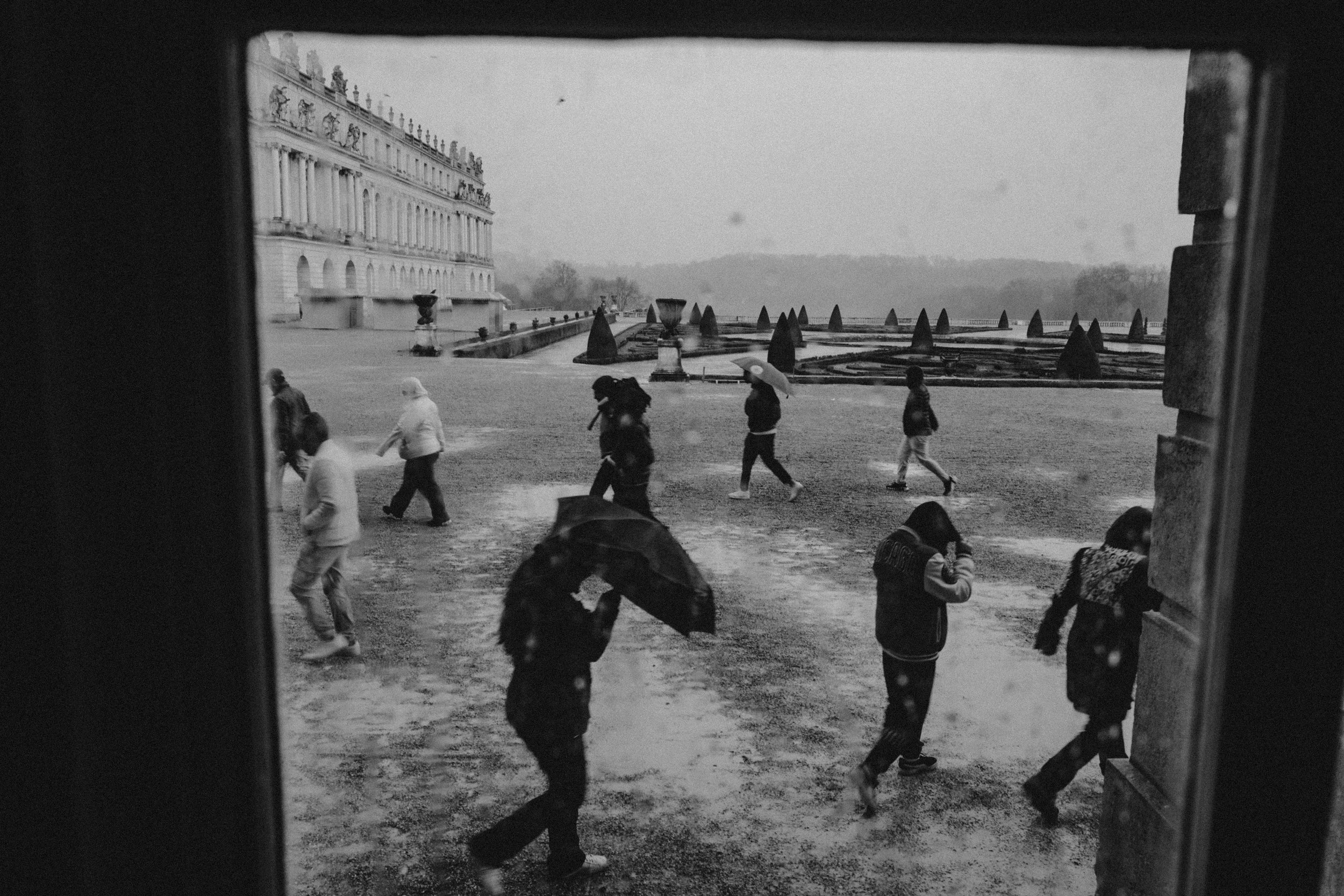 Black and white photo of people walking in the rain outside of the Palace of Versailles with multiple balconies and sculptures, viewed through a window frame with raindrops on it.