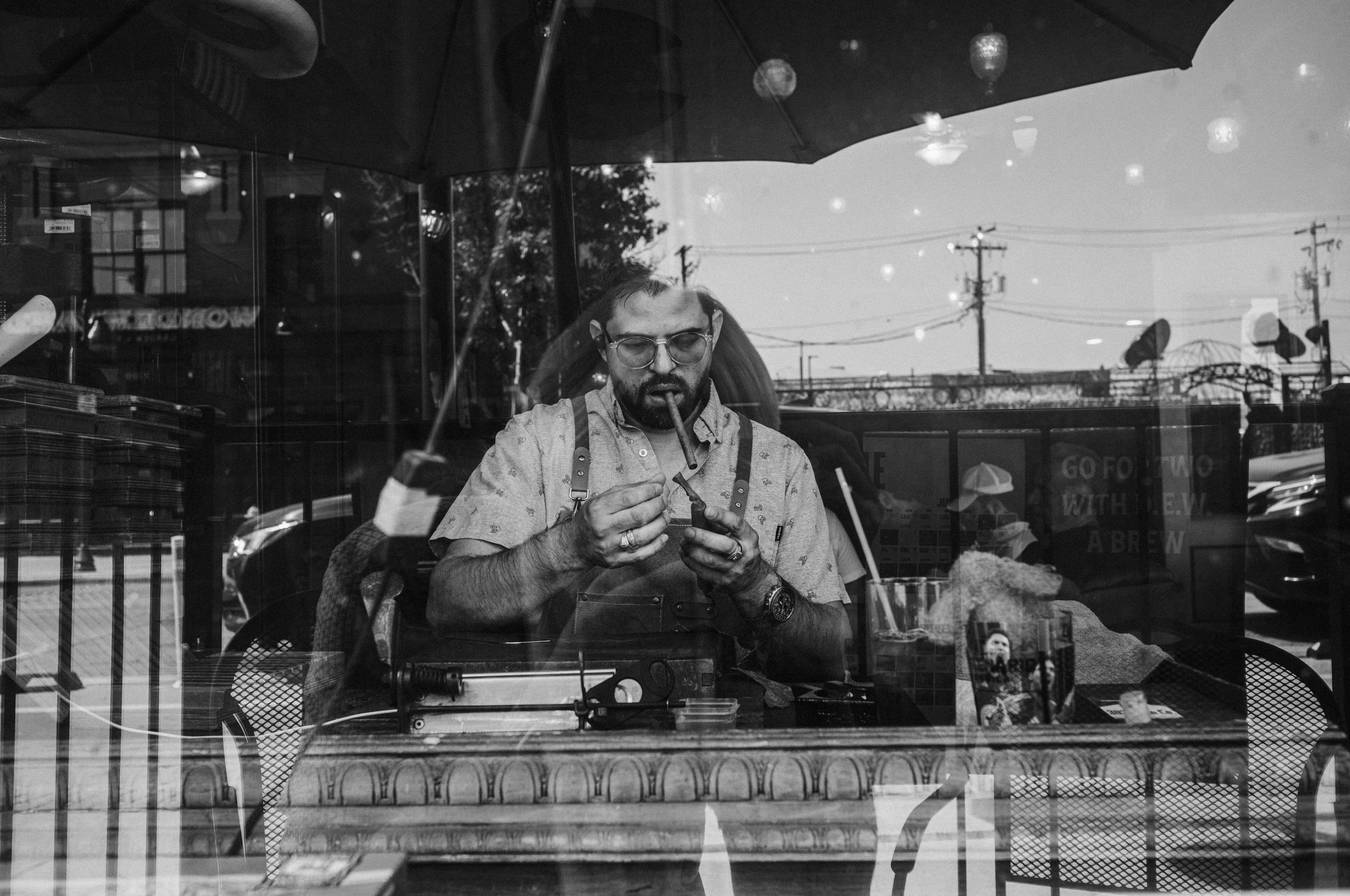 A man with glasses and a beard sitting at a table inside a restaurant or cafe, seen through a window, with reflections of streetlights and power lines outside. He is looking at his phone with a cigar in his mouth, and a straw in his drink on the tabl