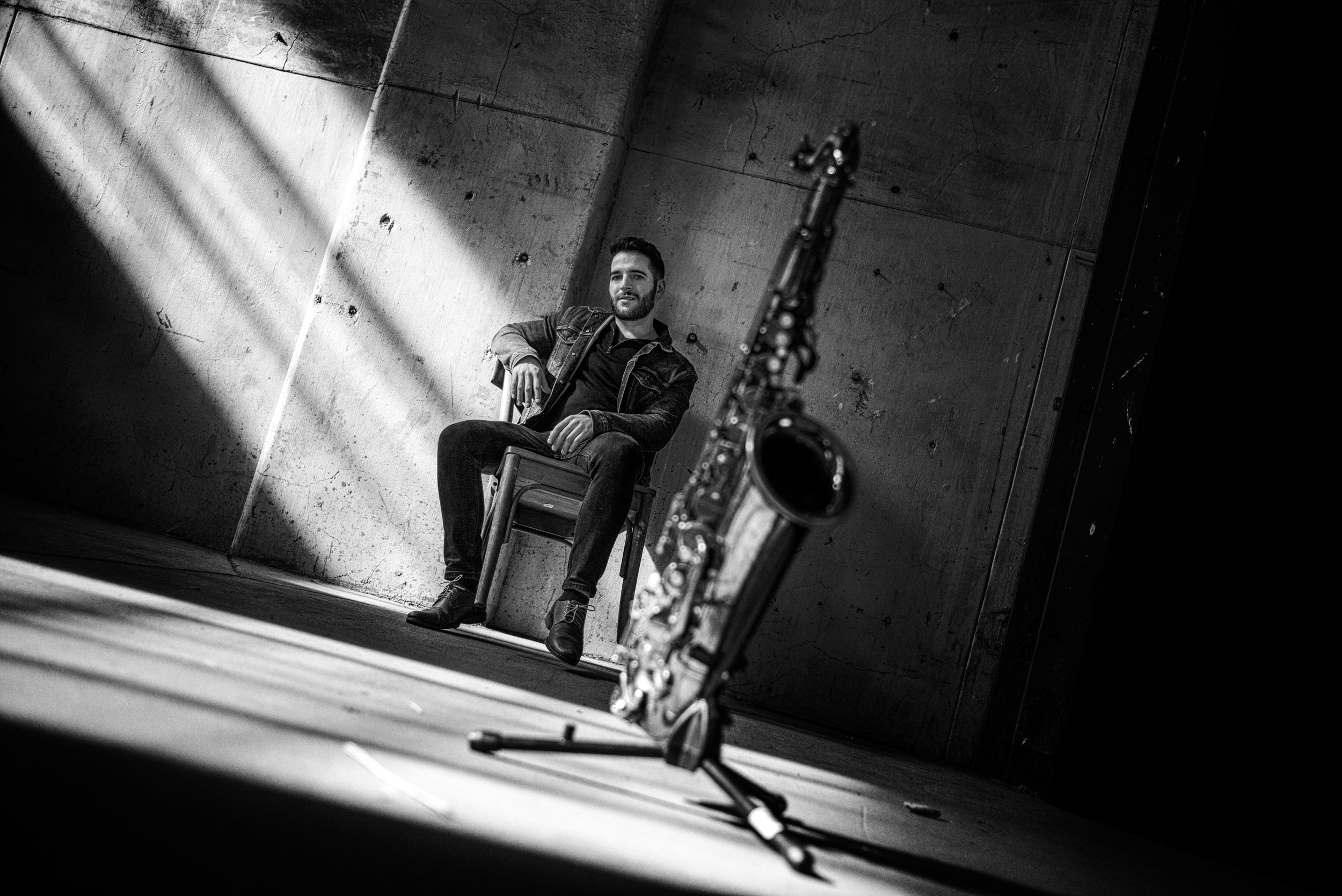 A black-and-white photo of a man sitting on a chair against a concrete wall, looking towards the camera with a slight smile. A saxophone is positioned in the foreground, slightly out of focus. Light streams in from the left, creating shadows on the w
