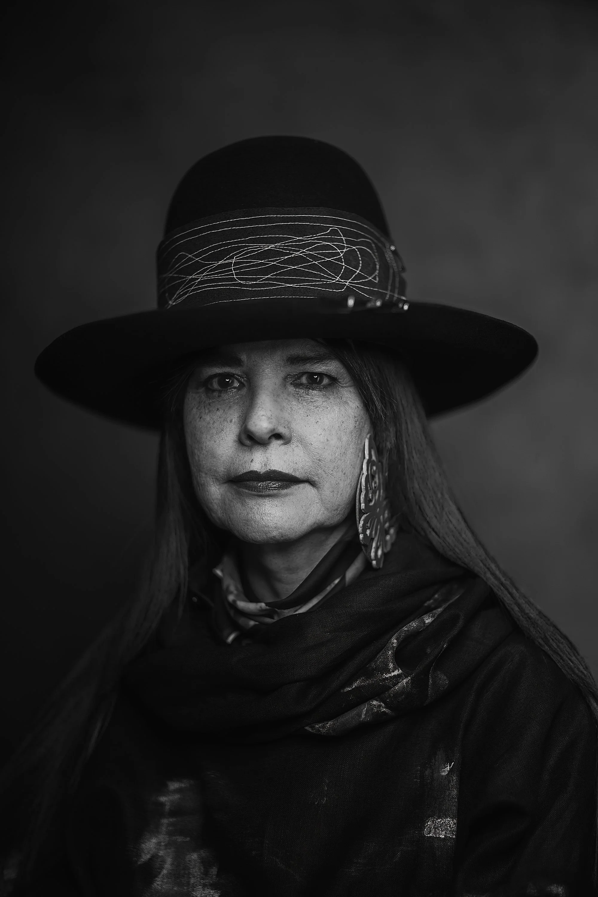 Black and white portrait of a woman wearing a large hat with scribble-like decorations, earrings, a scarf, and a dark cloak, looking directly at the camera against a dark background.