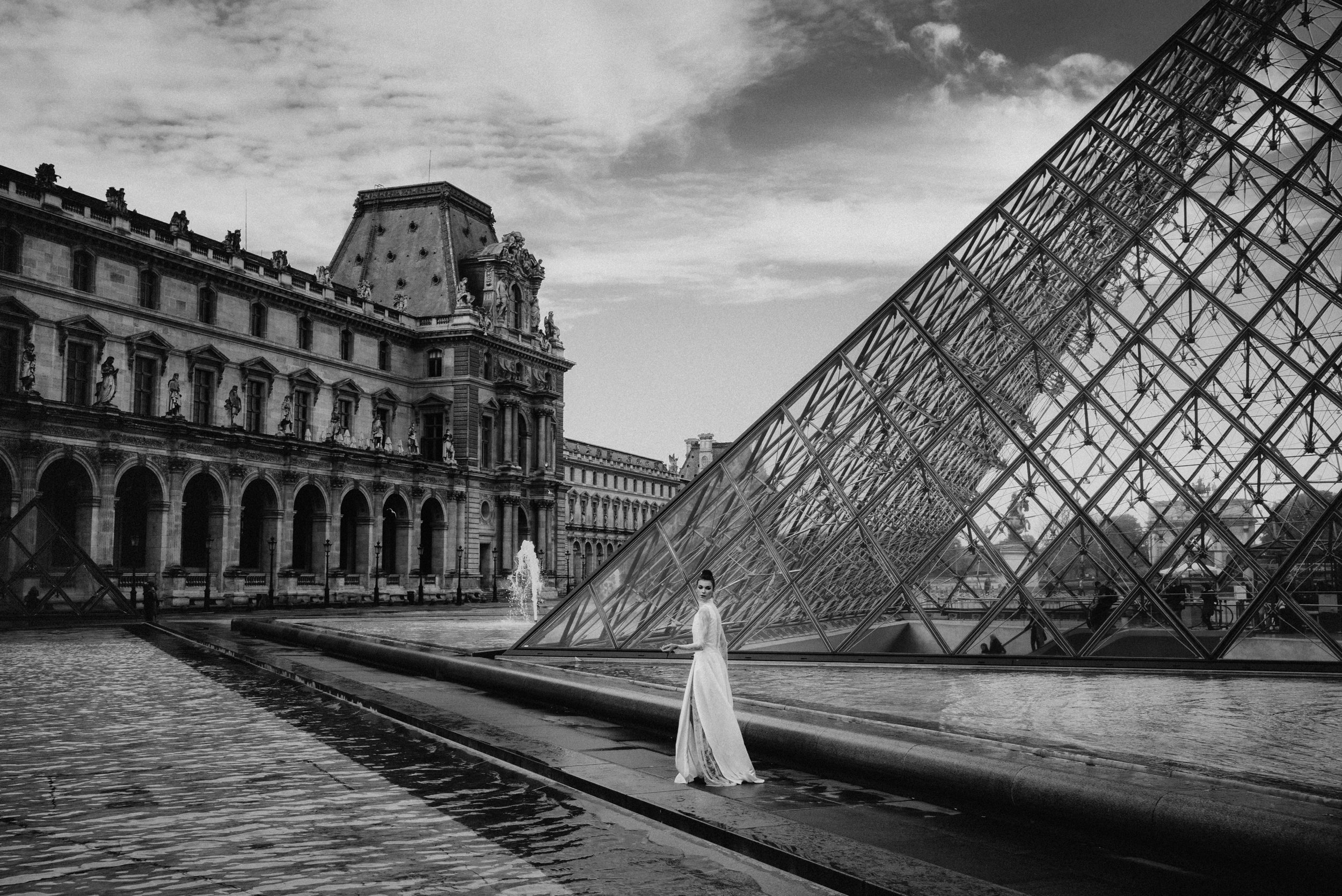 Black and white photo of the Louvre Museum in Paris with a glass pyramid sculpture in the foreground, and a woman in a white dress standing by the water fountain.