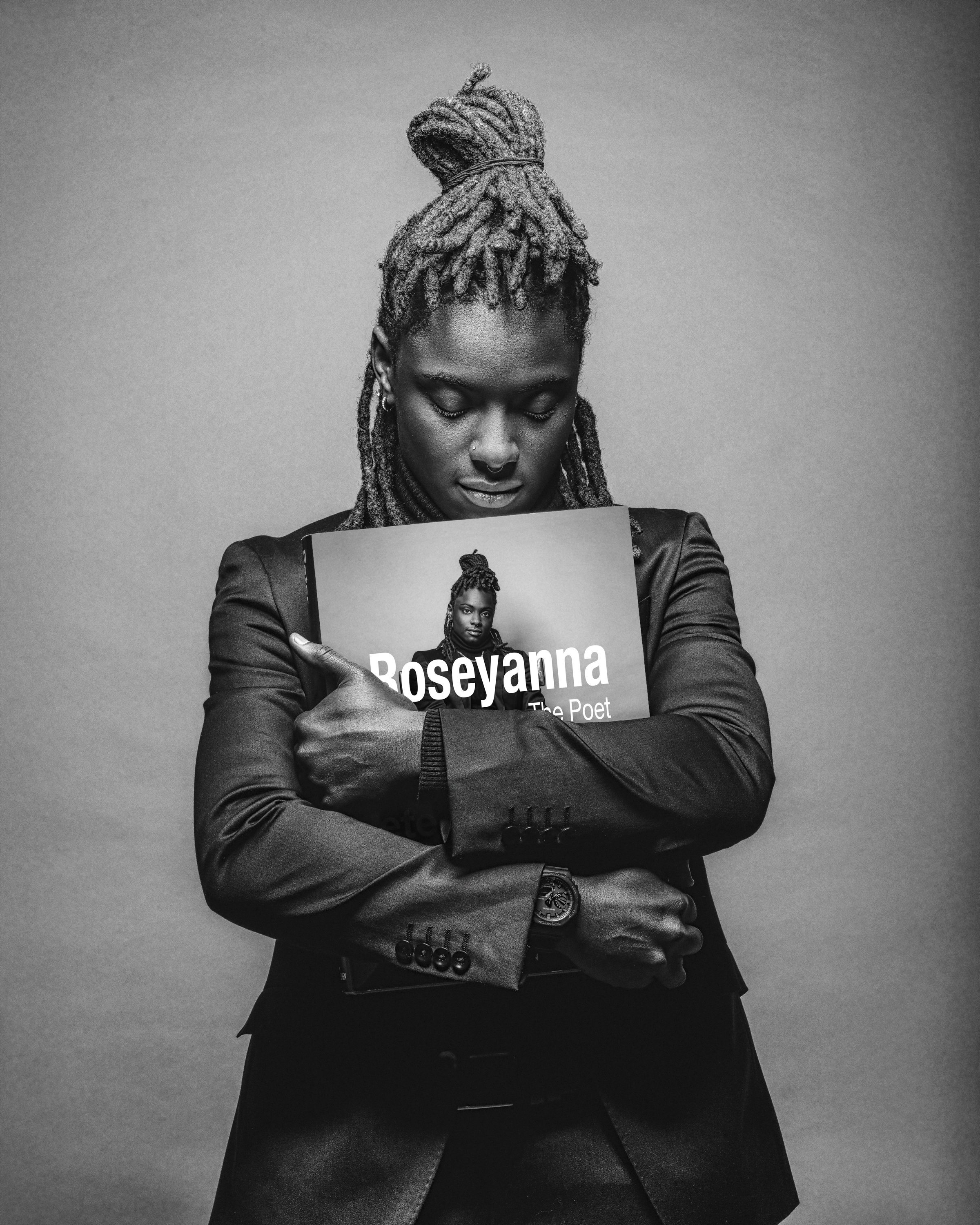 A woman with dreadlocks styled in a high bun, holding a book with her eyes closed, wearing a dark blazer and a watch, in black and white.