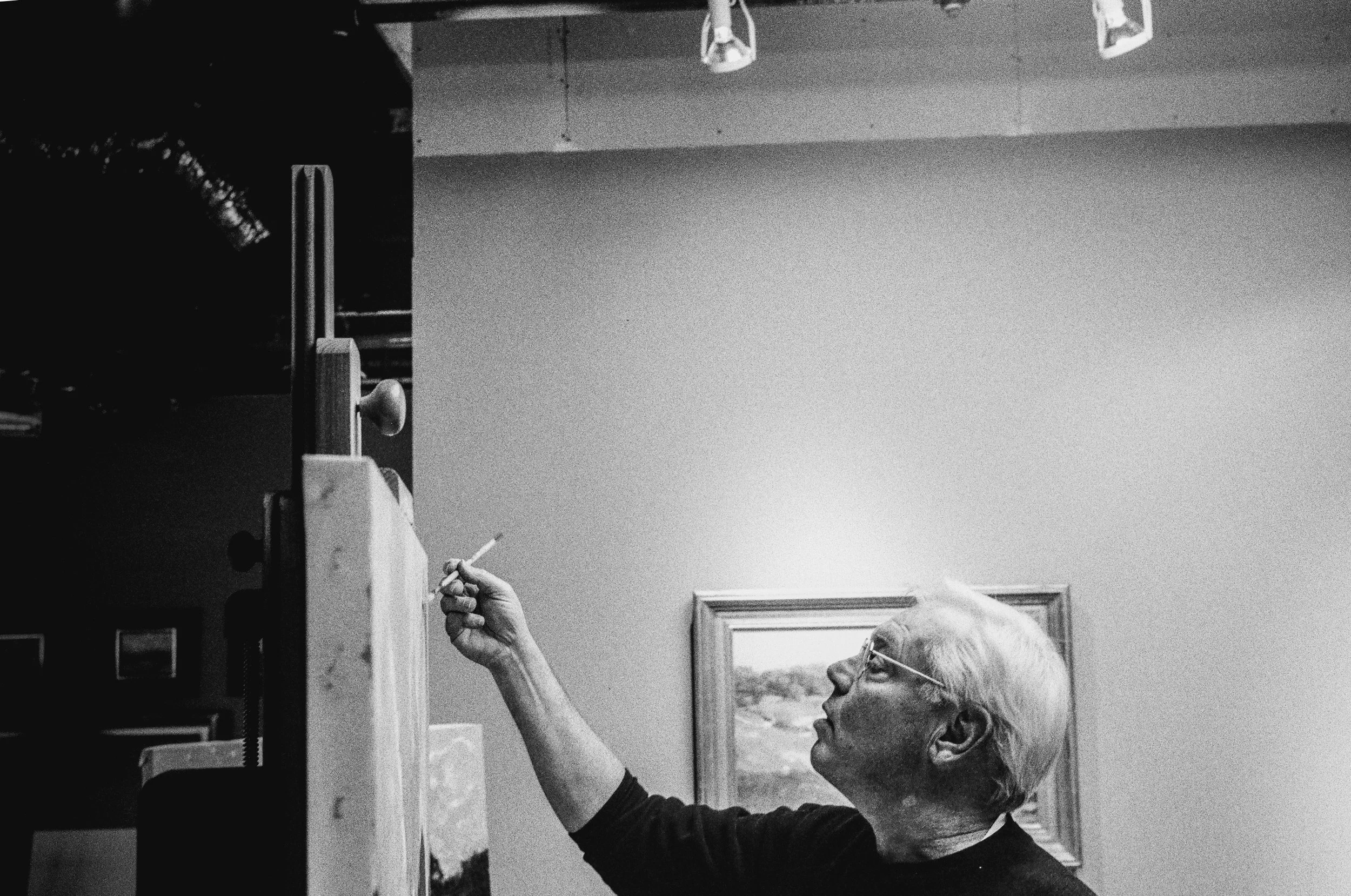 An elderly man with glasses and short hair painting or restoring a picture on a wall, using a brush, with the focus on her hand and the art piece.