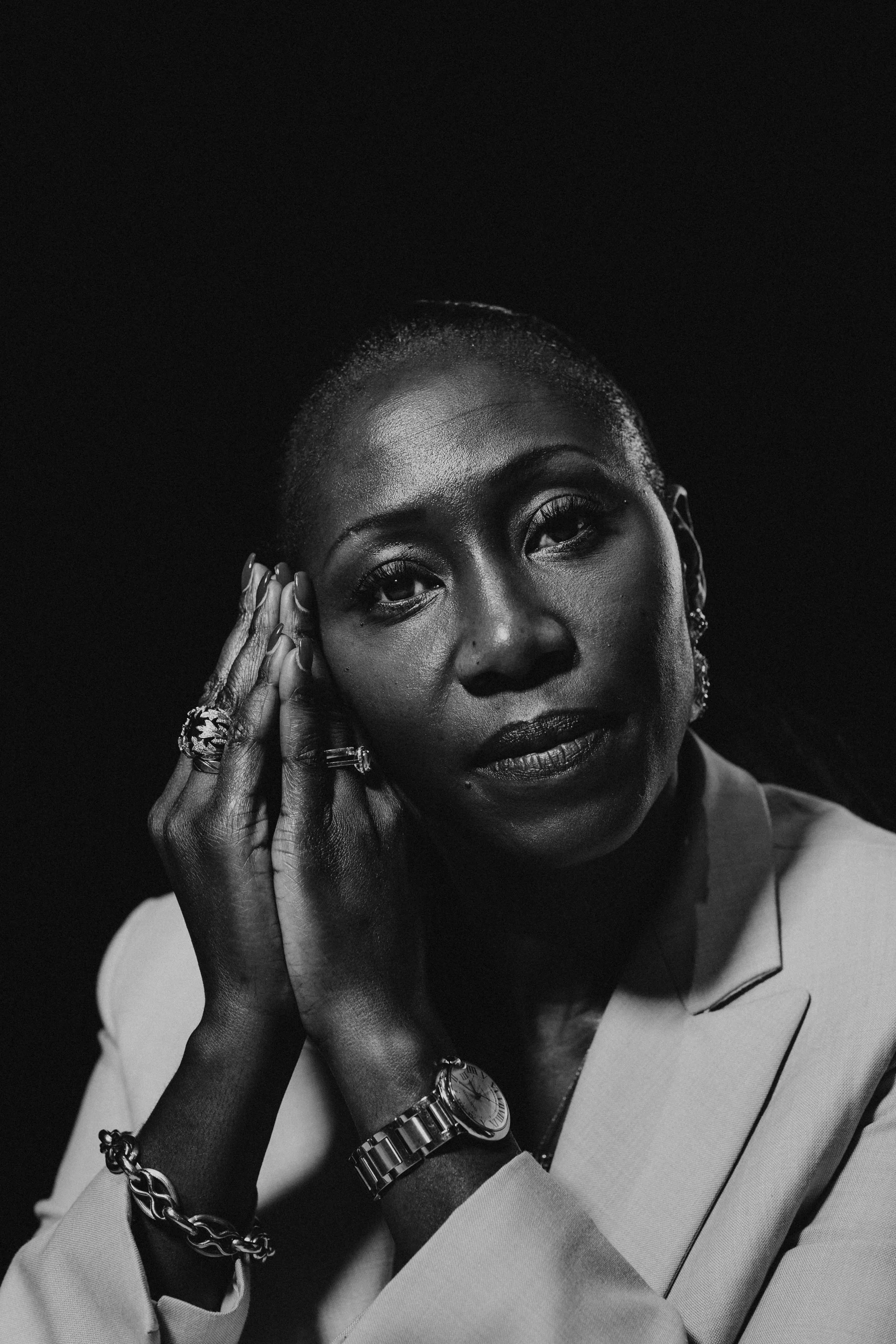 Black and white portrait of a woman with short hair, wearing jewelry including rings, a watch, bracelets, and earrings, with one hand touching her face.