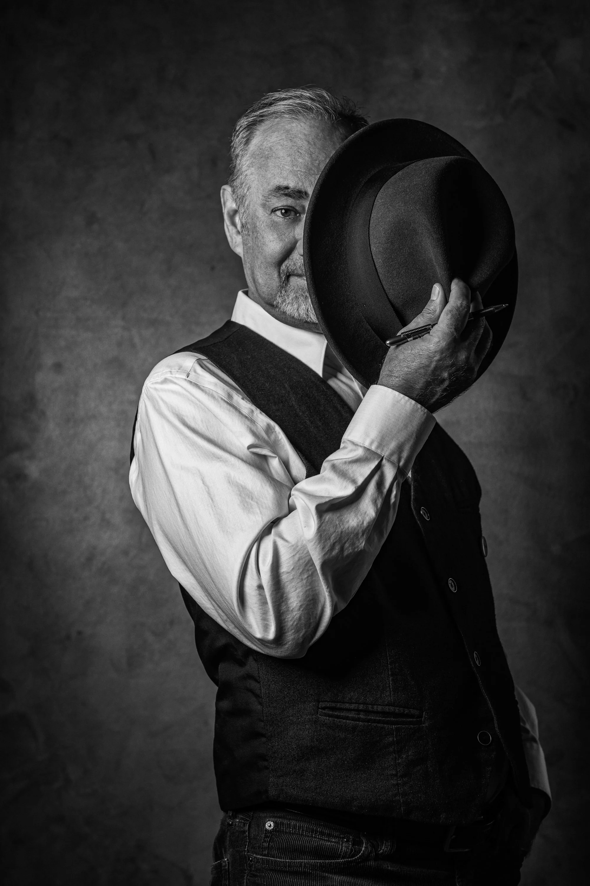 A black and white photo of a man holding a hat in front of his face, with one eye visible.