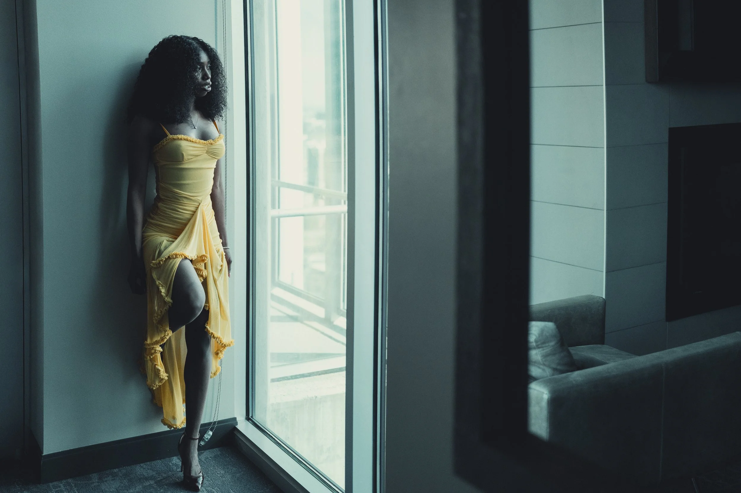 A woman with curly hair wearing a yellow dress and silver heels stands by a glass window, looking outside in a modern indoor space with a gray chair and tiled wall.