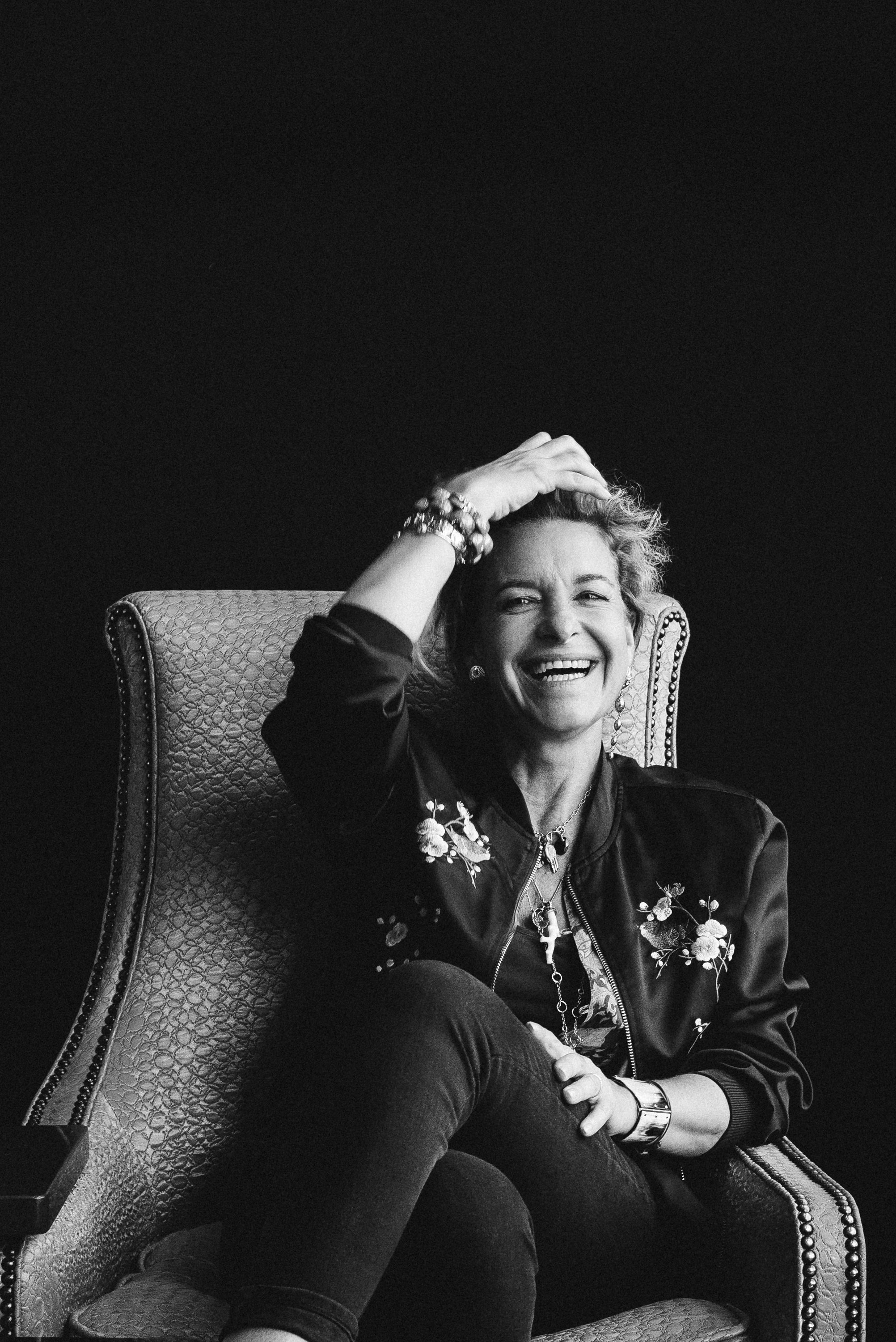 A woman with curly hair, smiling, sitting in a high-backed, patterned armchair. She is wearing a dark jacket with floral embroidery, a wristwatch, and bracelets, with her left hand resting on her knee. Her right hand is on her head, touching her hair