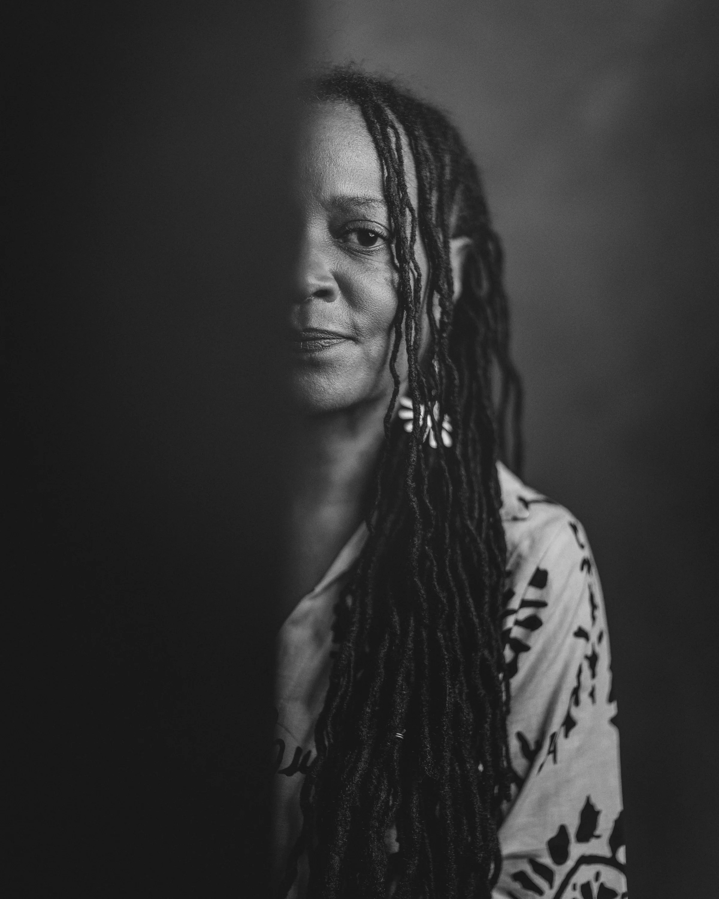 Black and white photo of a woman with dreadlocks and earrings, with a partially obscured face, wearing a patterned top.