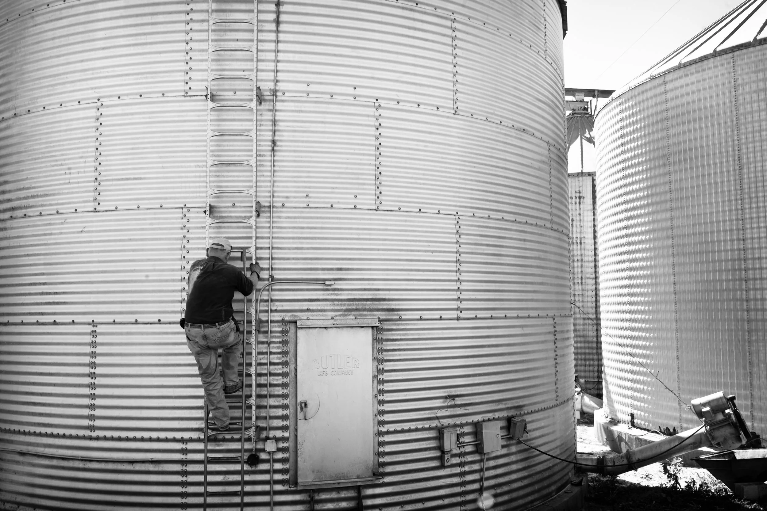 A farmer  standing on a ladder working on the exterior of a large metal grain silo, with another silo partially visible on the right.
