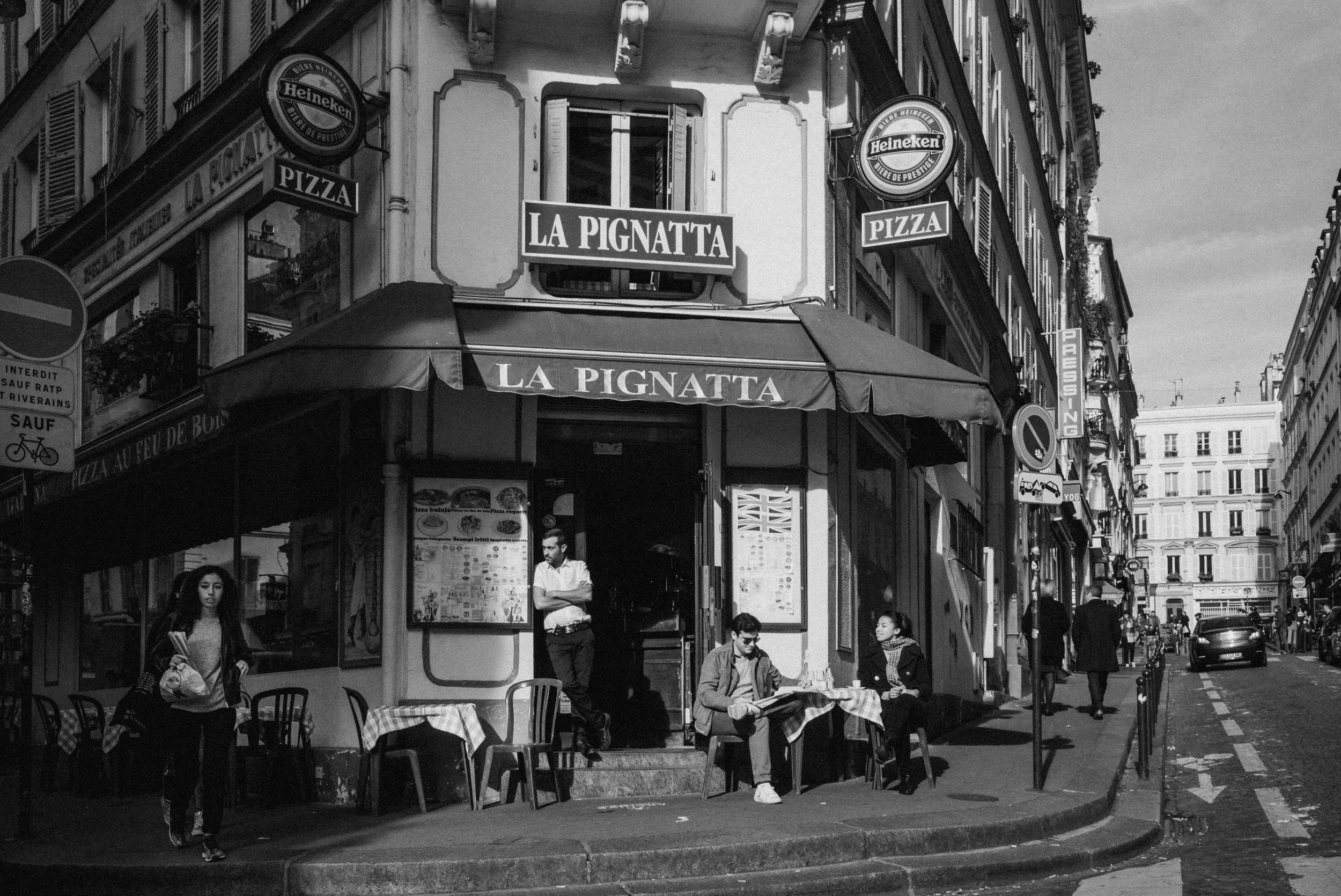 A street scene in Paris with a restaurant named La Pignatta and outdoor seating, pedestrians walking along the sidewalk, and buildings with typical European architecture.