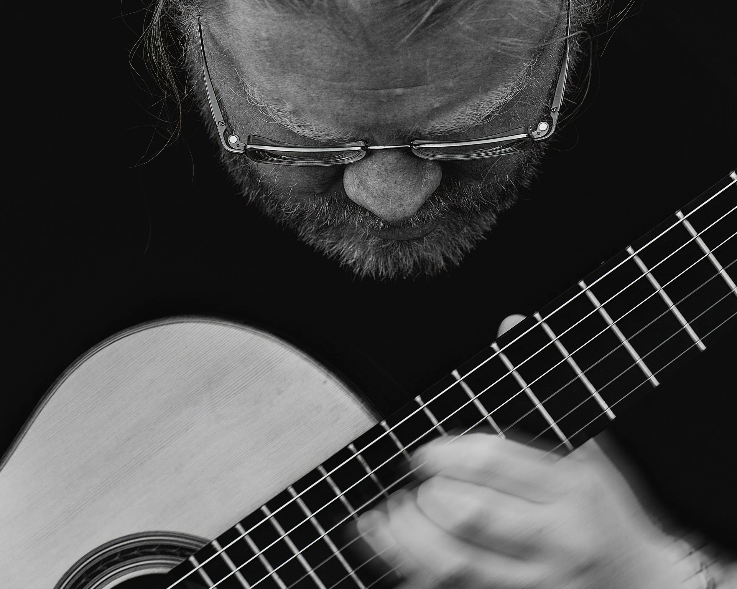 Close-up of a man with glasses playing an acoustic guitar, black and white photo.