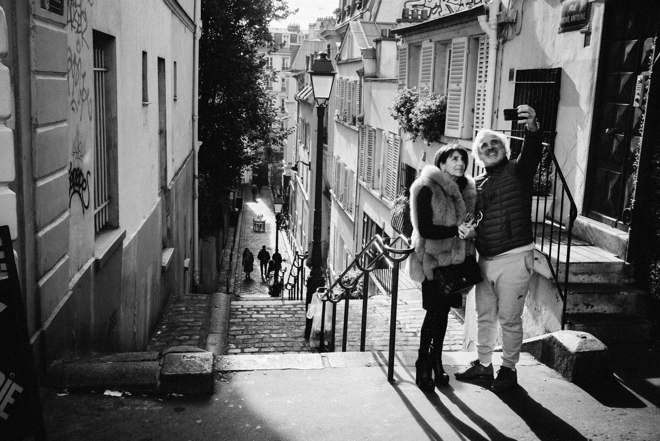 A black and white photo of a man and woman taking a selfie on a staircase in a Parisian neighborhood with cobblestone street and classic European buildings with shuttered windows.
