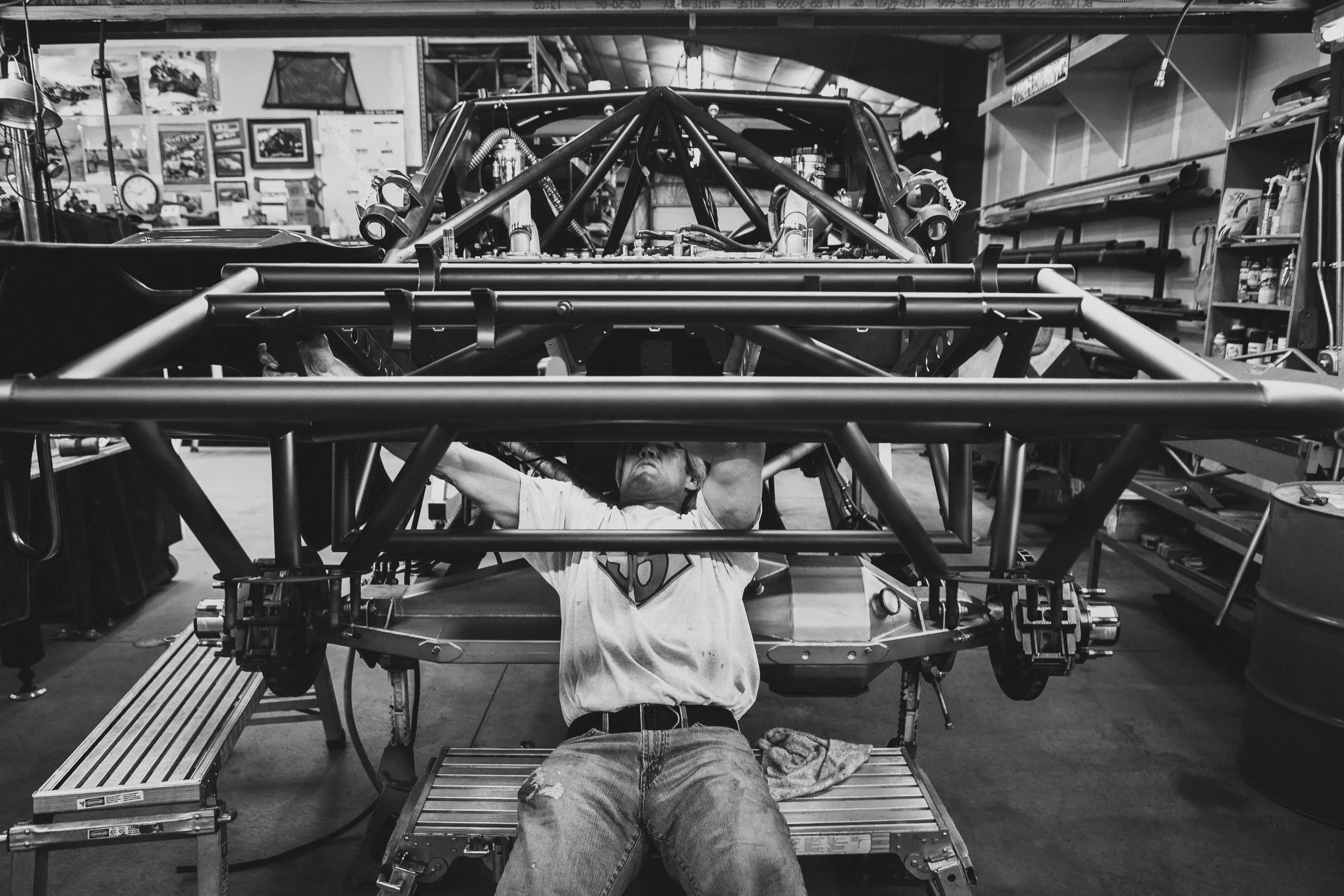 Person working underneath a race car frame in a garage or workshop environment.