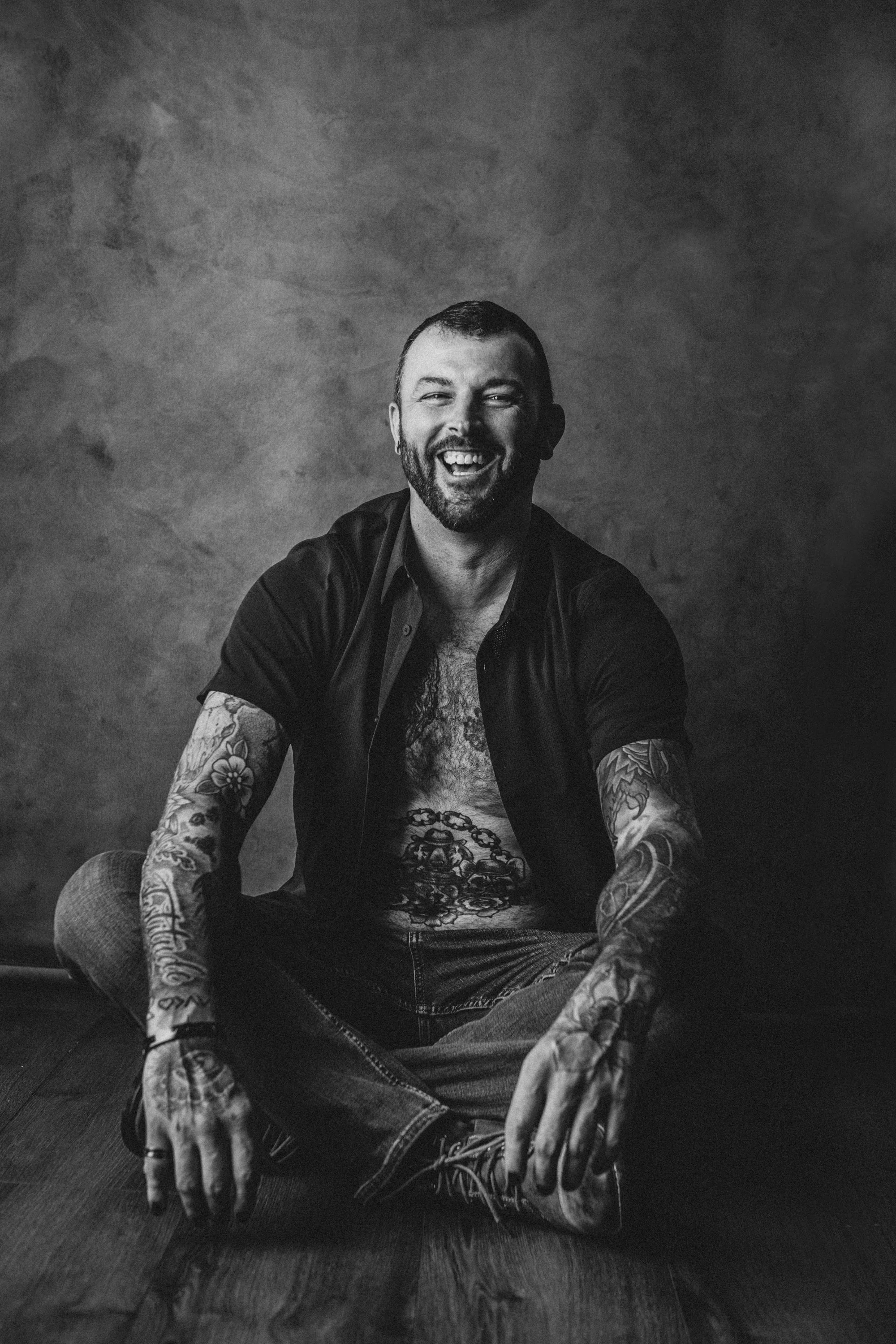 Black and white photo of a smiling man with tattoos, sitting cross-legged on the floor against a plain wall.