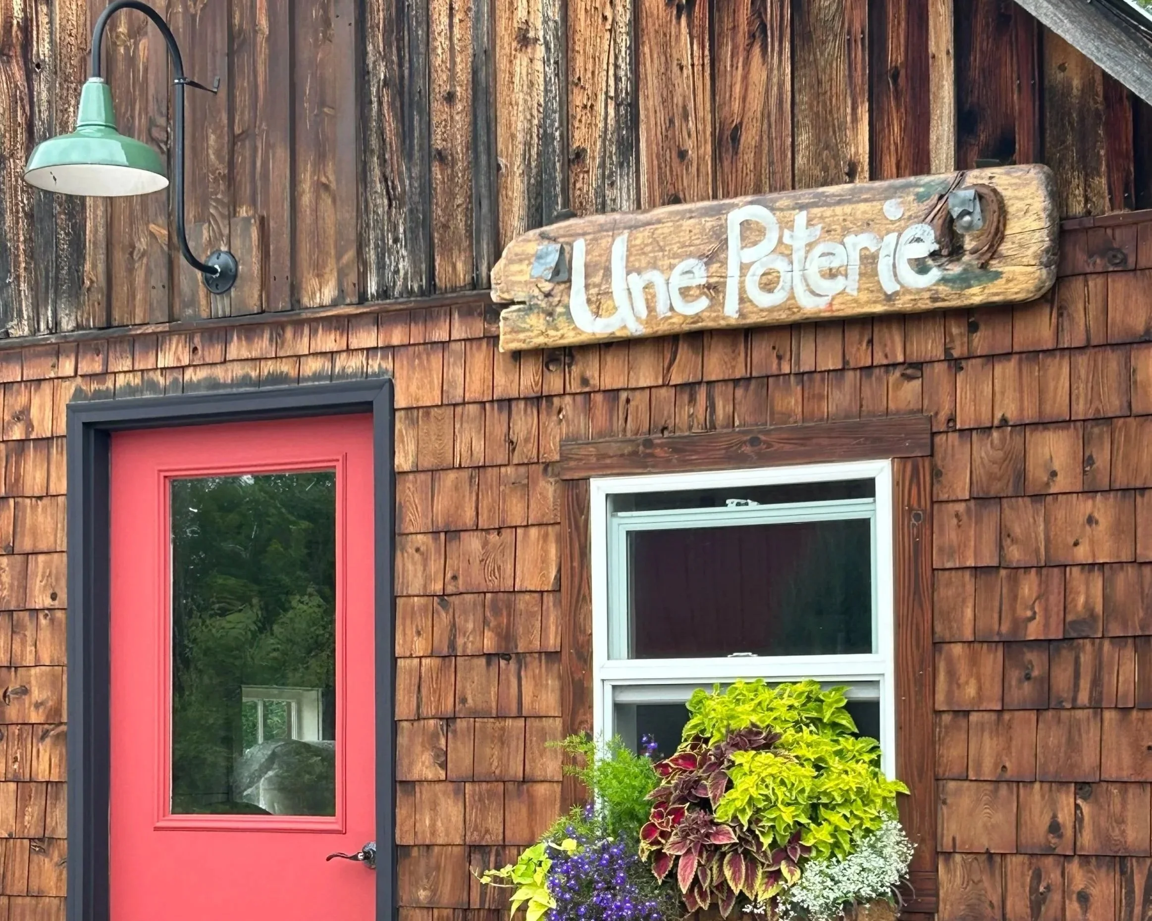 Rustic wooden pottery studio with a red door, Une Poterie sign and colorful flowers.