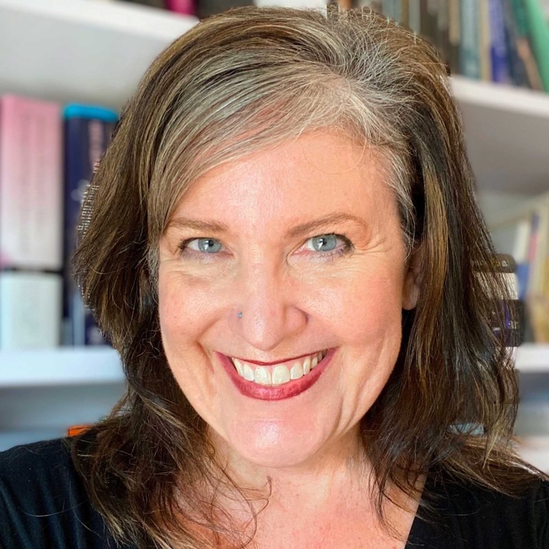 Smiling middle-aged woman with brown and gray hair, blue eyes, and red lipstick in front of a bookshelf