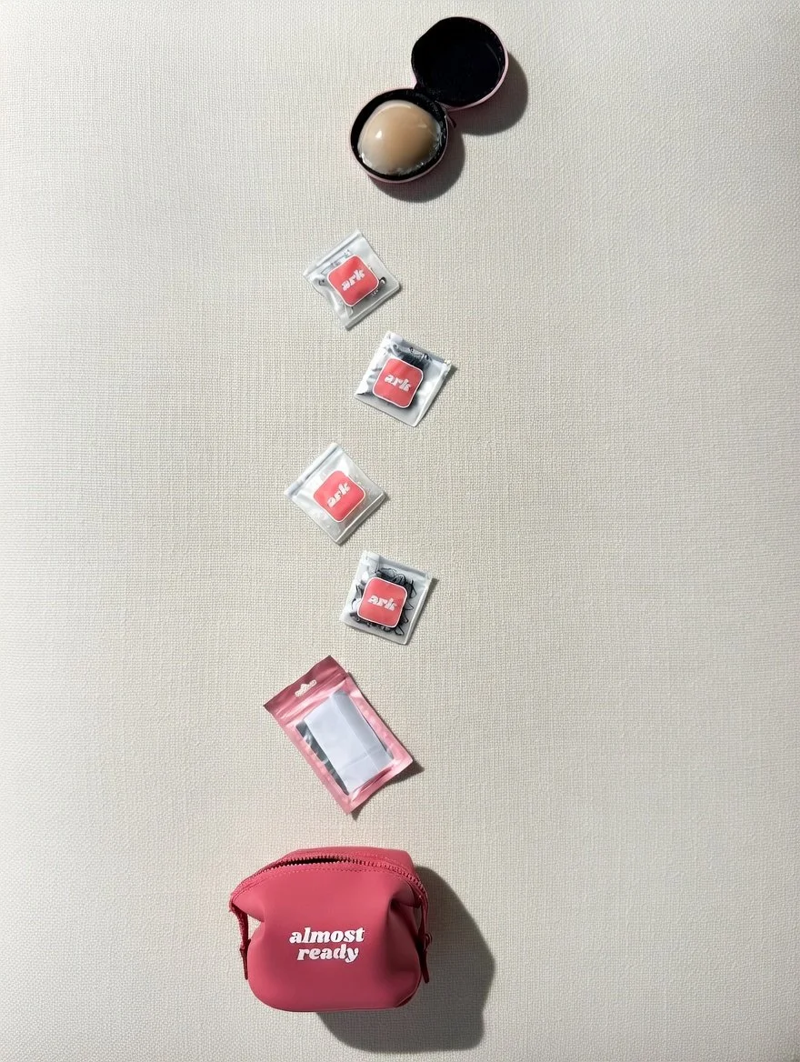 A pink pouch with the text 'almost ready' surrounded by small individually packed items and two open containers, all placed on a light-colored surface. women gift idea. travel pouch. travel kit.