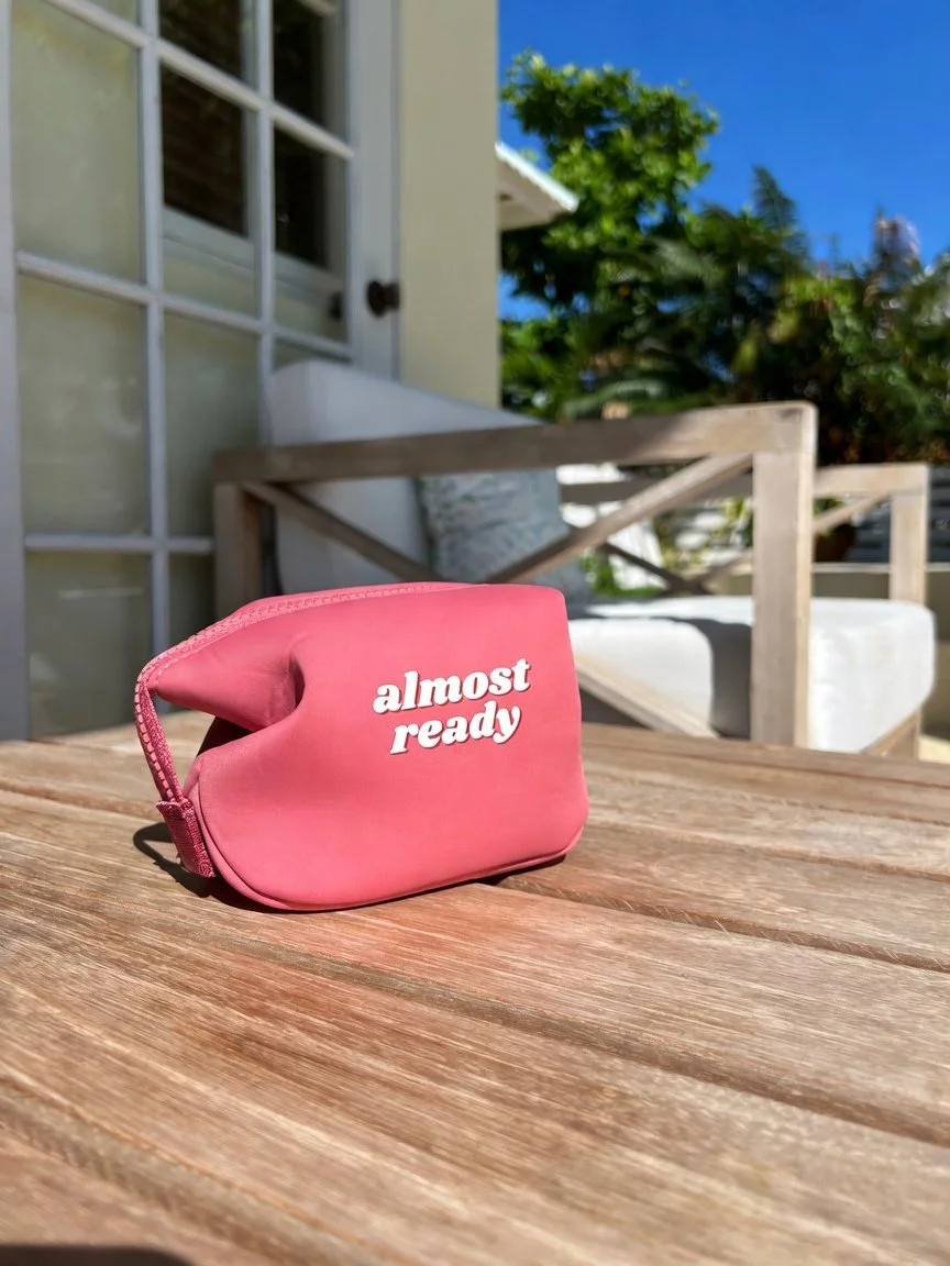 A small pink pouch with the text "almost ready" in white letters sitting on a wooden outdoor table under sunlight, with a background of greenery, a house door, and a white outdoor seat.  women gift idea. travel pouch. travel kit.