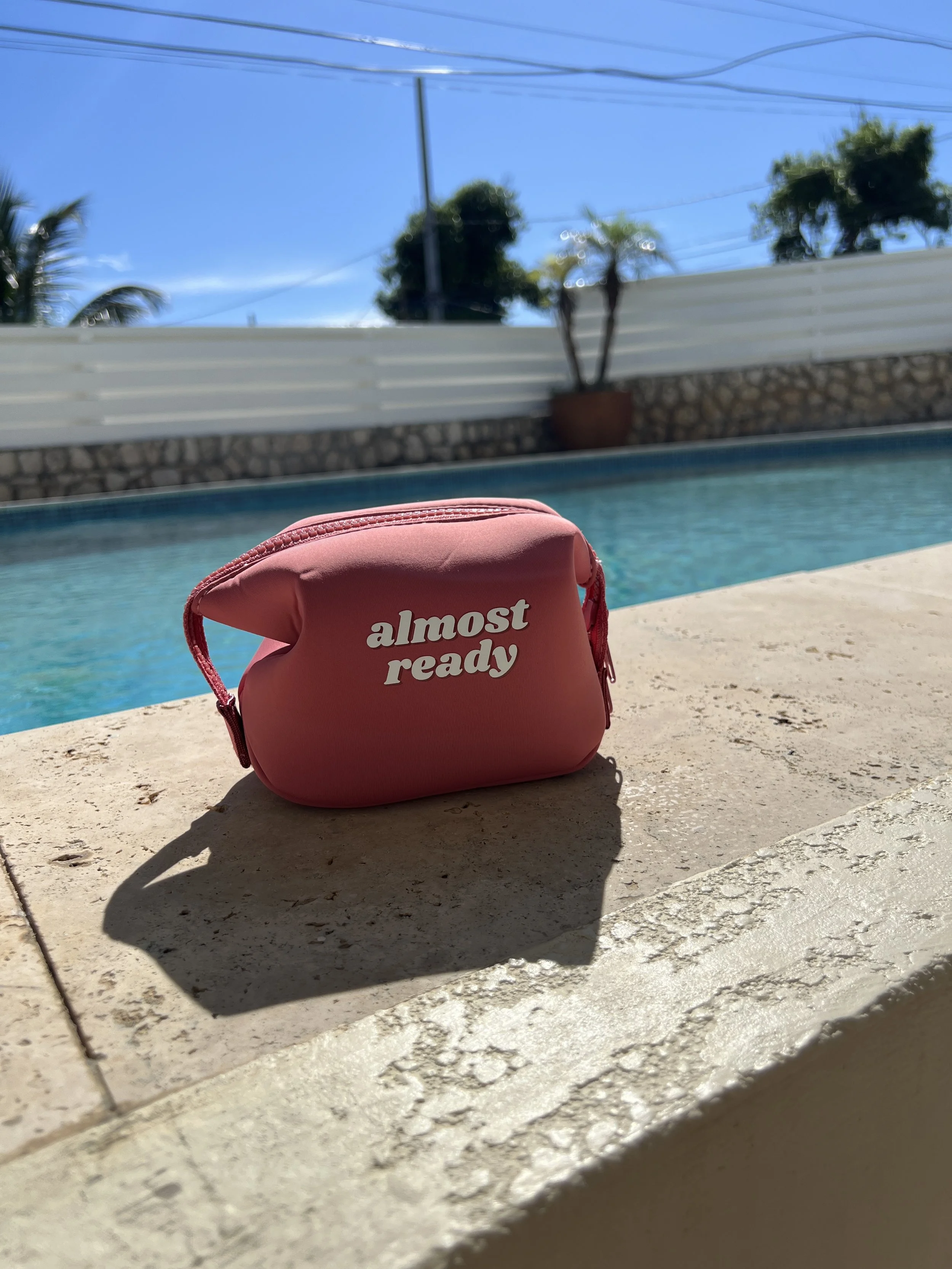 Pink gift pouch with white text that says 'almost ready' next to a swimming pool during daytime.  women gift idea. travel pouch. travel kit.