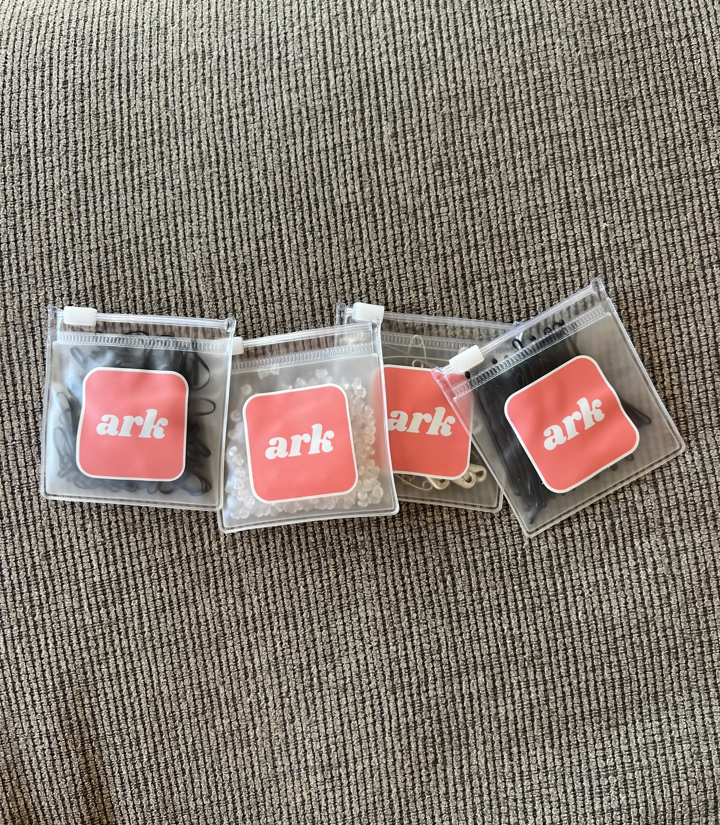 Four small plastic bags with black hair clips inside, placed on a brown textured carpet. Each bag has a red label with the word 'ark' in white.