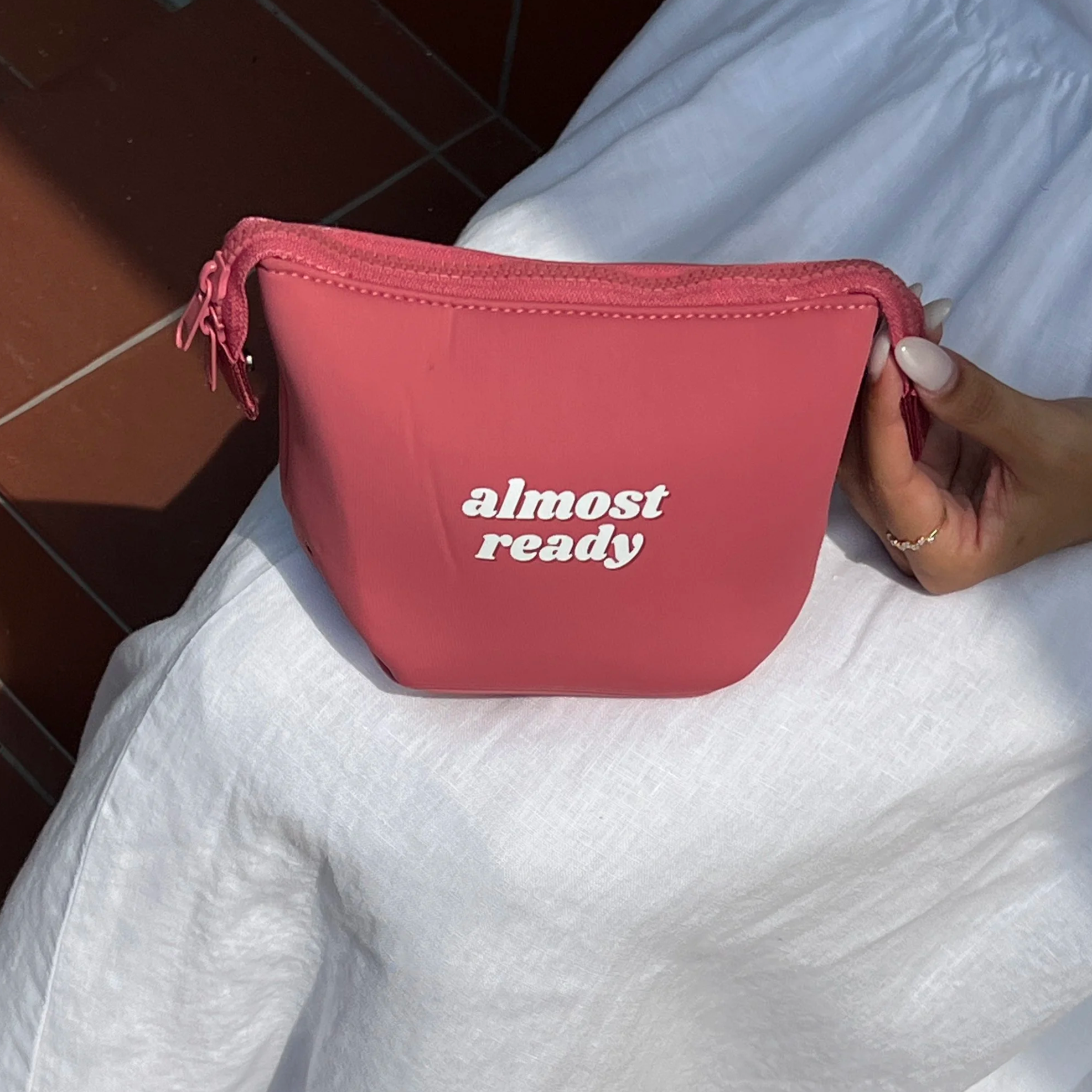 A pink zippered pouch with white text reading 'almost ready' held in a person's hand, with a white or light-colored fabric and a dark tiled surface in the background.  women gift idea. travel pouch. travel kit.