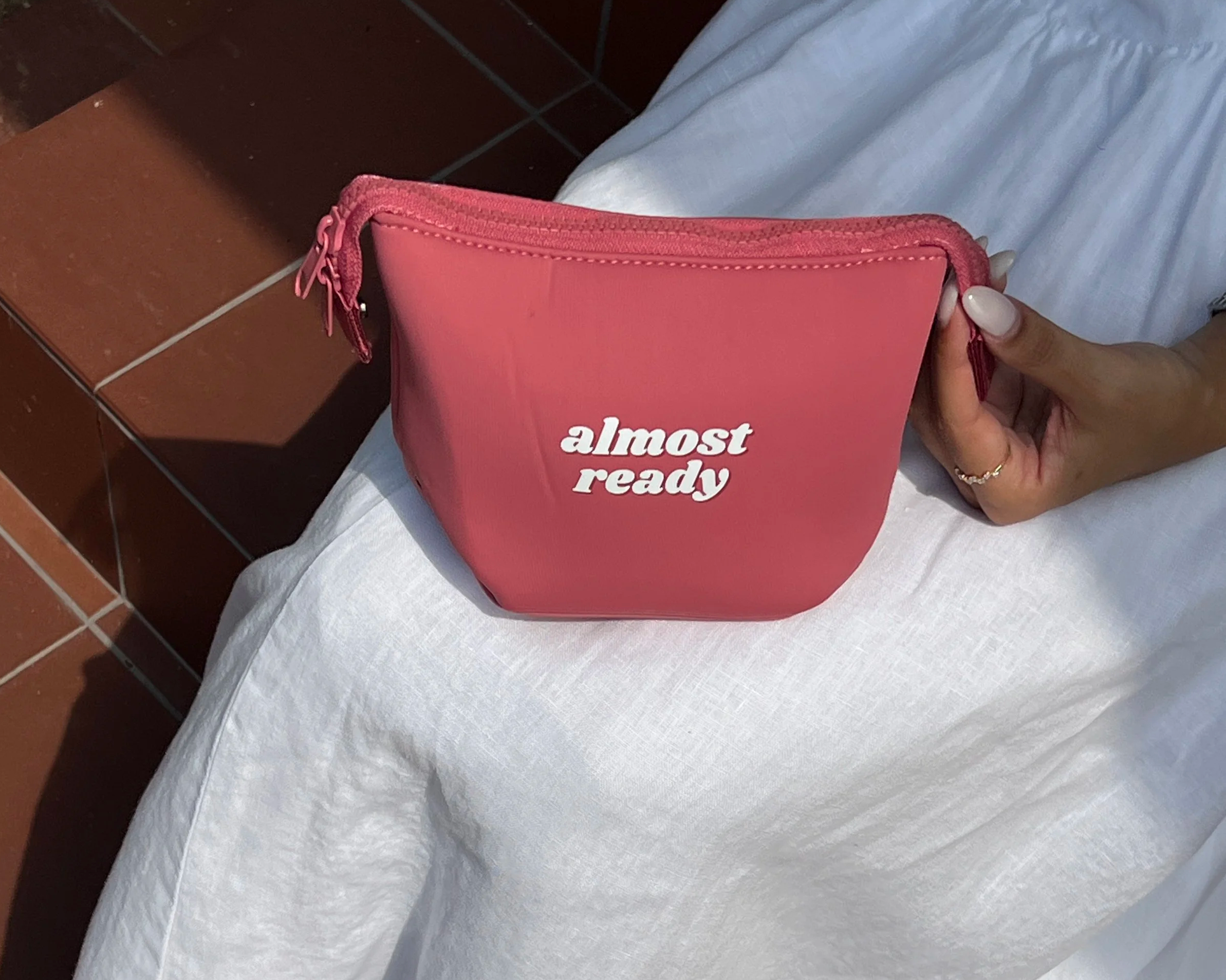 A person holding a pink wallet or pouch with the text 'almost ready' written on it, sitting on a white cloth with a brown tiled surface nearby.