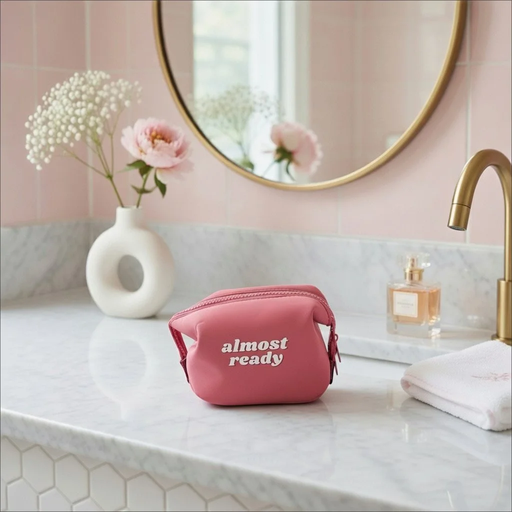 A pink toiletry bag with white text that says "almost ready" sits on a white marble bathroom counter. Behind it, a white vase with pink flowers and white baby's breath, a mirror, a gold faucet, a bottle of liquid soap, and a white towel are visible.