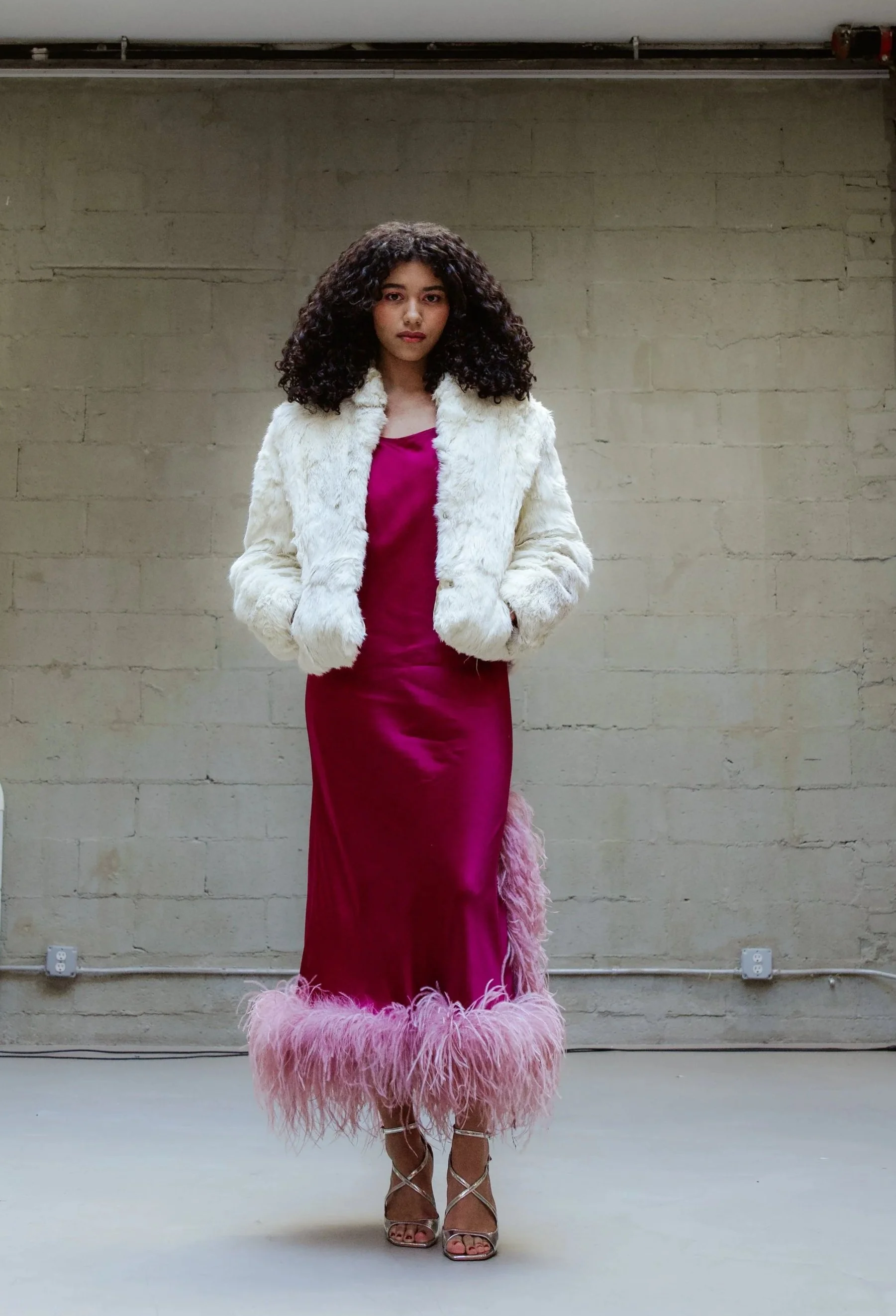 A woman standing against a plain beige brick wall, wearing a fuchsia satin dress with a matching feather trim, a white faux fur jacket, and high-heeled strappy sandals.