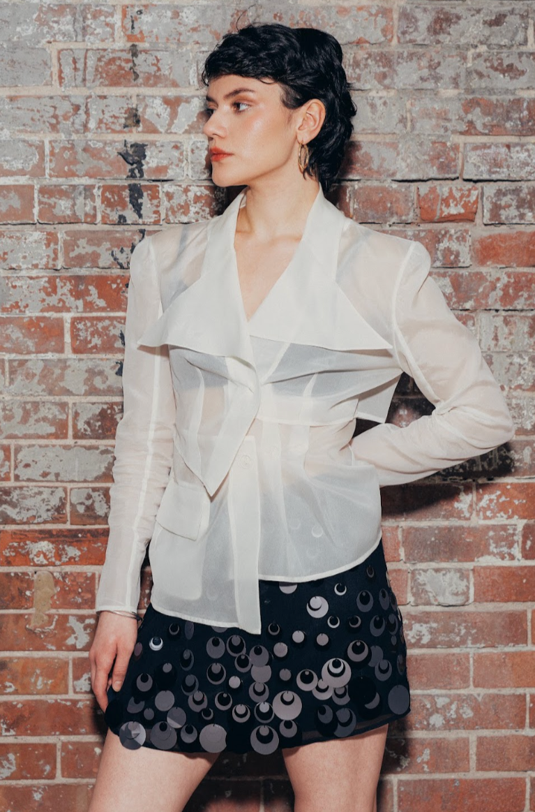 A woman with short black hair and hoop earrings stands against a brick wall, wearing a sheer white blouse and a black skirt with circular sequins.