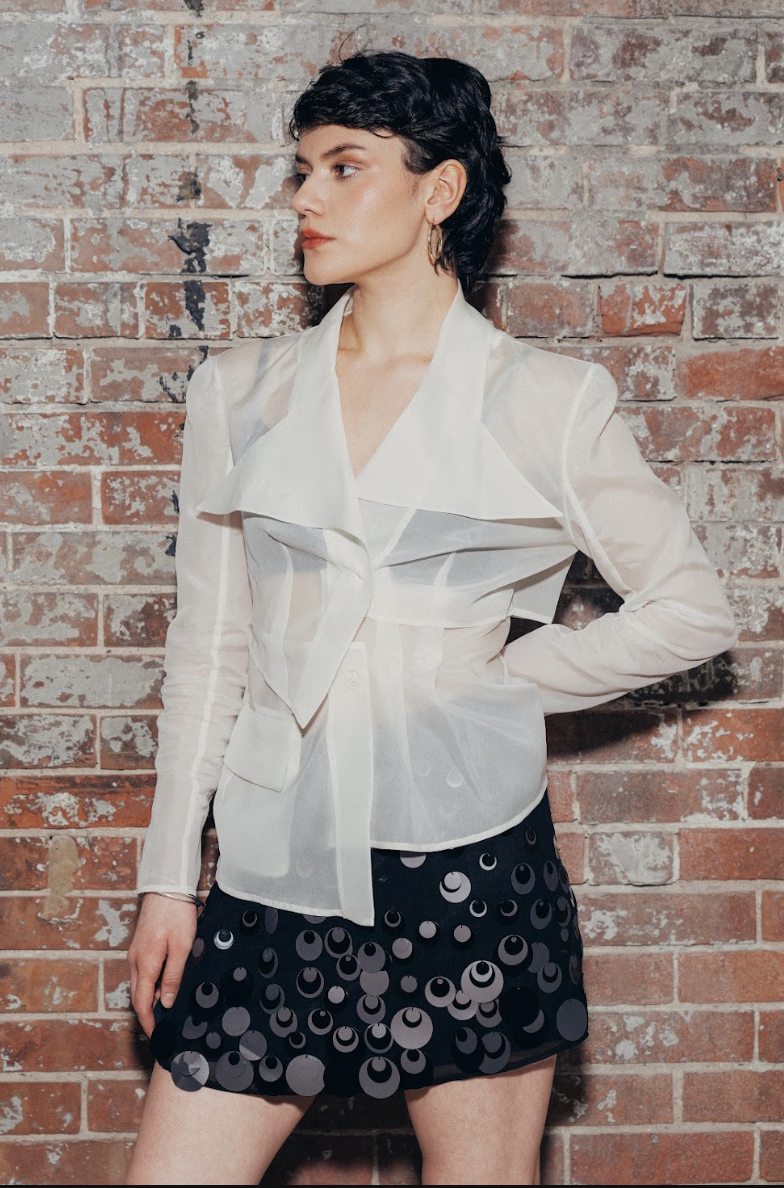 A woman with short black hair and hoop earrings stands against a brick wall, wearing a sheer white blouse and a black skirt with circular sequins.