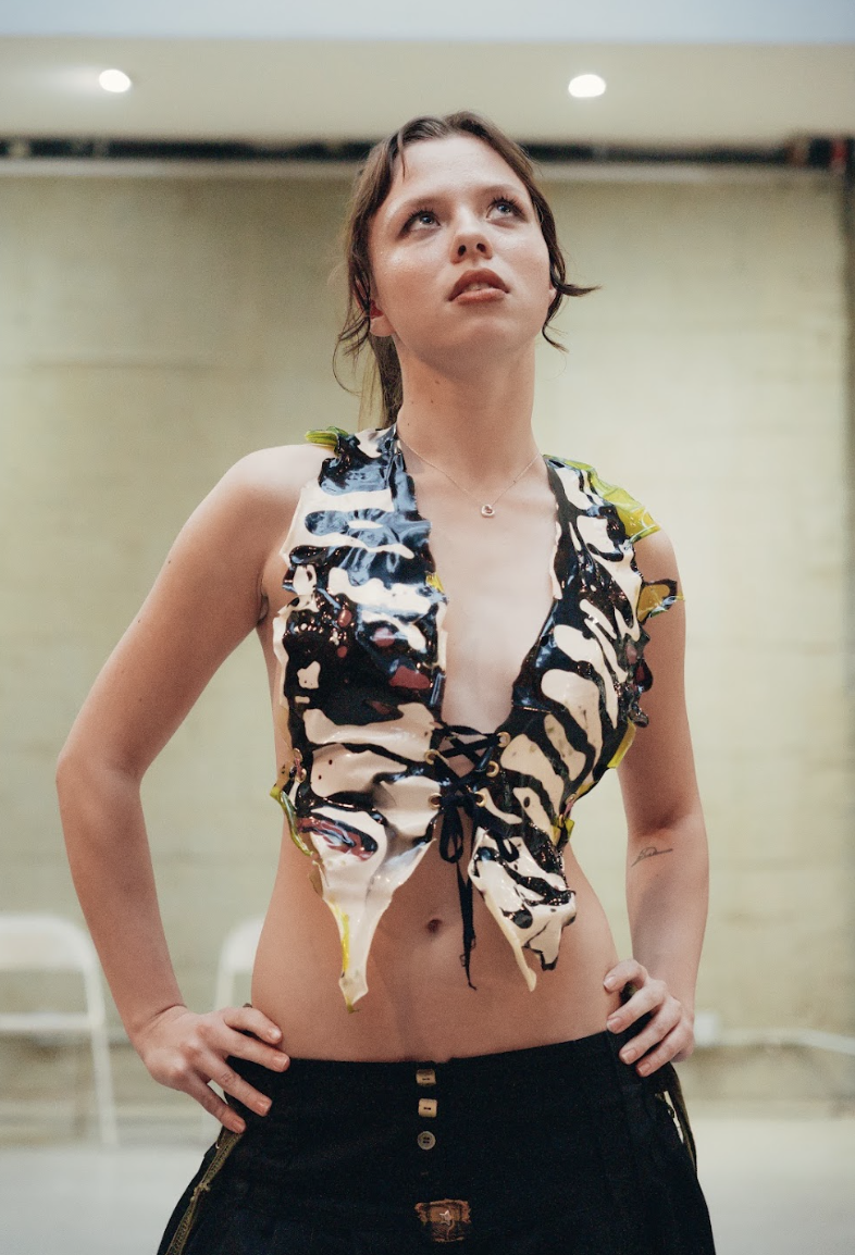 Young woman with short brown hair wearing a colorful, ruffled, sleeveless top with a lace-up front and dark jeans, standing with her hands on her hips in an indoor setting.