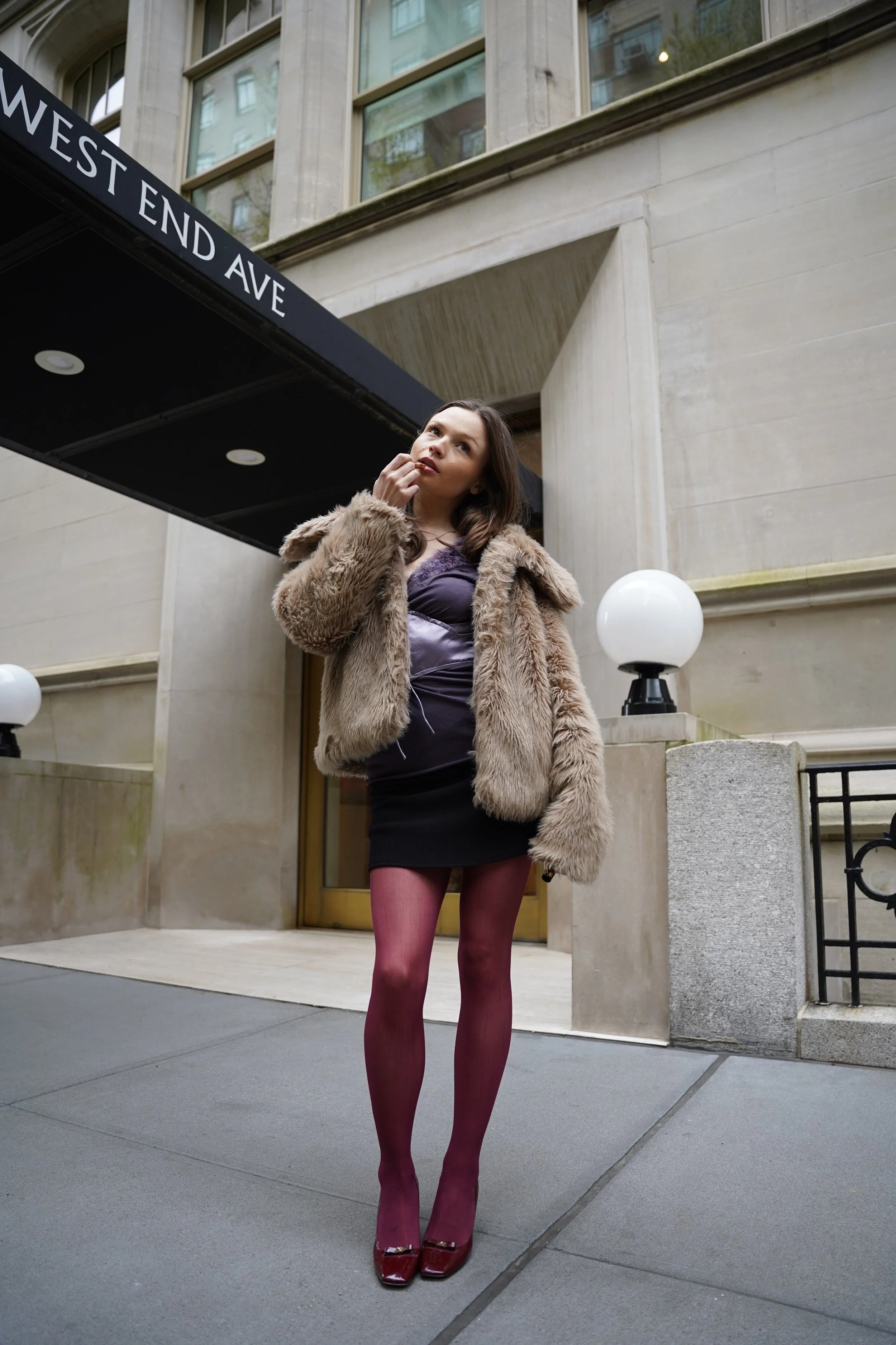 A young woman with dark hair standing on a city sidewalk outside a building with large windows and a black awning that reads "WEST END AVE." She is wearing a fur coat, purple dress, red tights, and red high-heeled shoes.