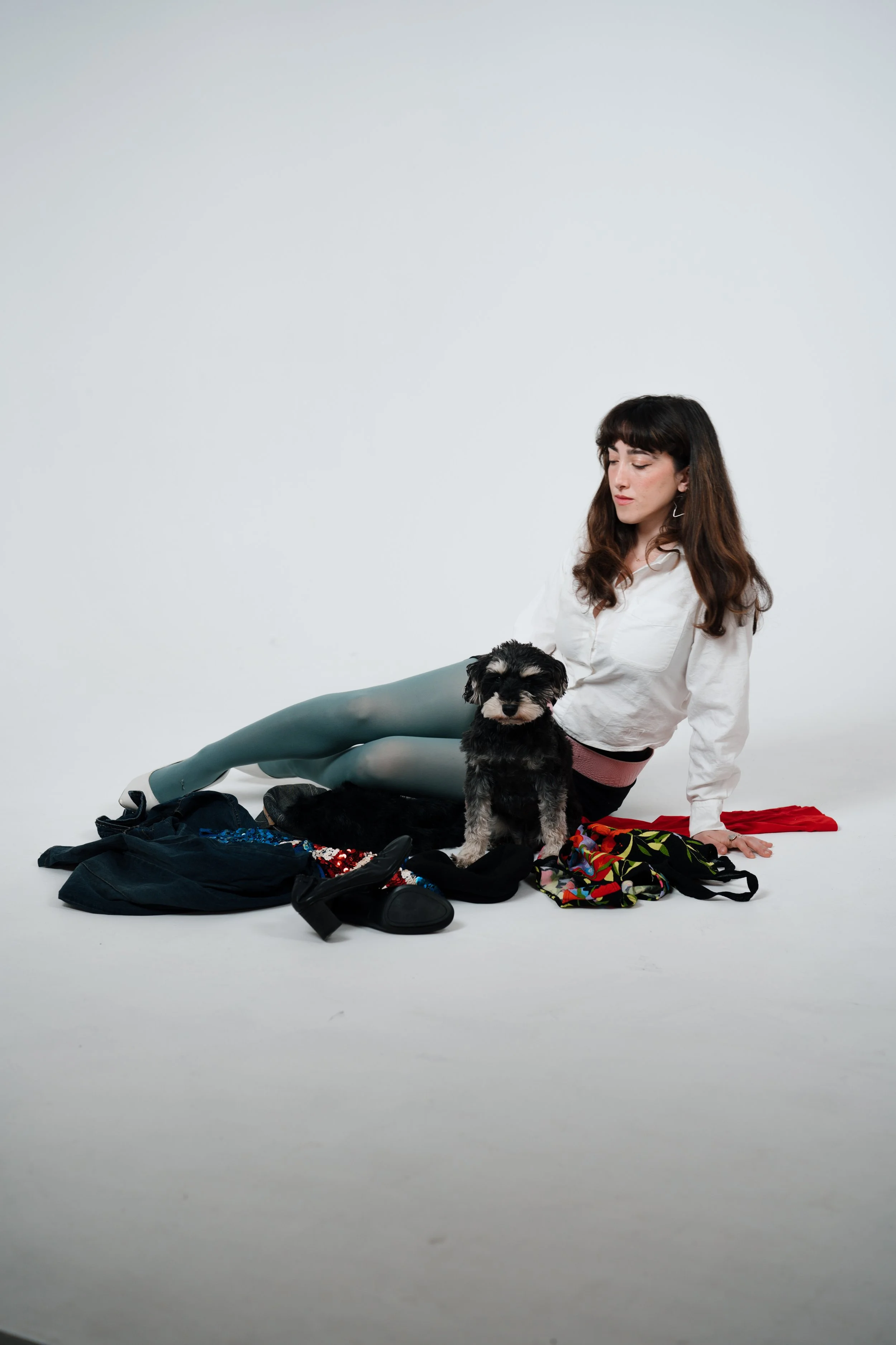 A woman with long dark hair wearing a white shirt and teal leggings sitting on the floor with scattered clothing and accessories around her, a small black and gray dog sitting in front of her, against a plain white background.