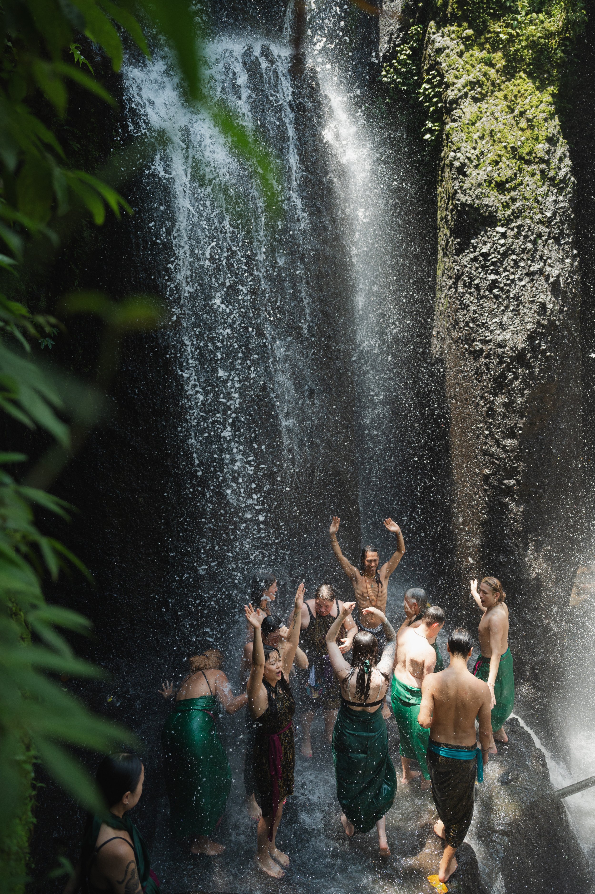 Group of people dancing beneath a tropical waterfall surrounded by lush greenery