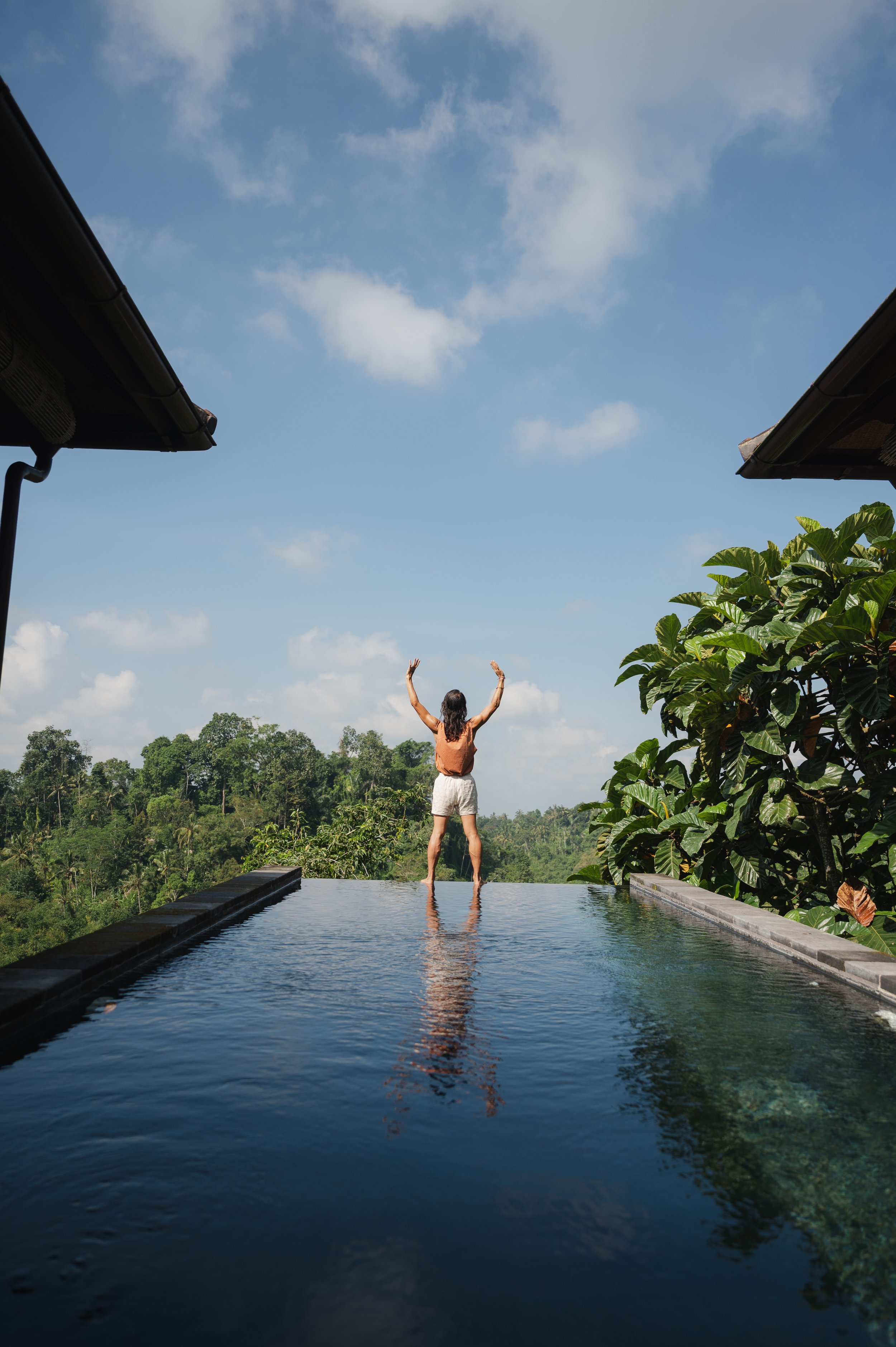 Person with raised arms standing in an infinity pool overlooking a lush tropical landscape, framed by villa roofs and blue sky.