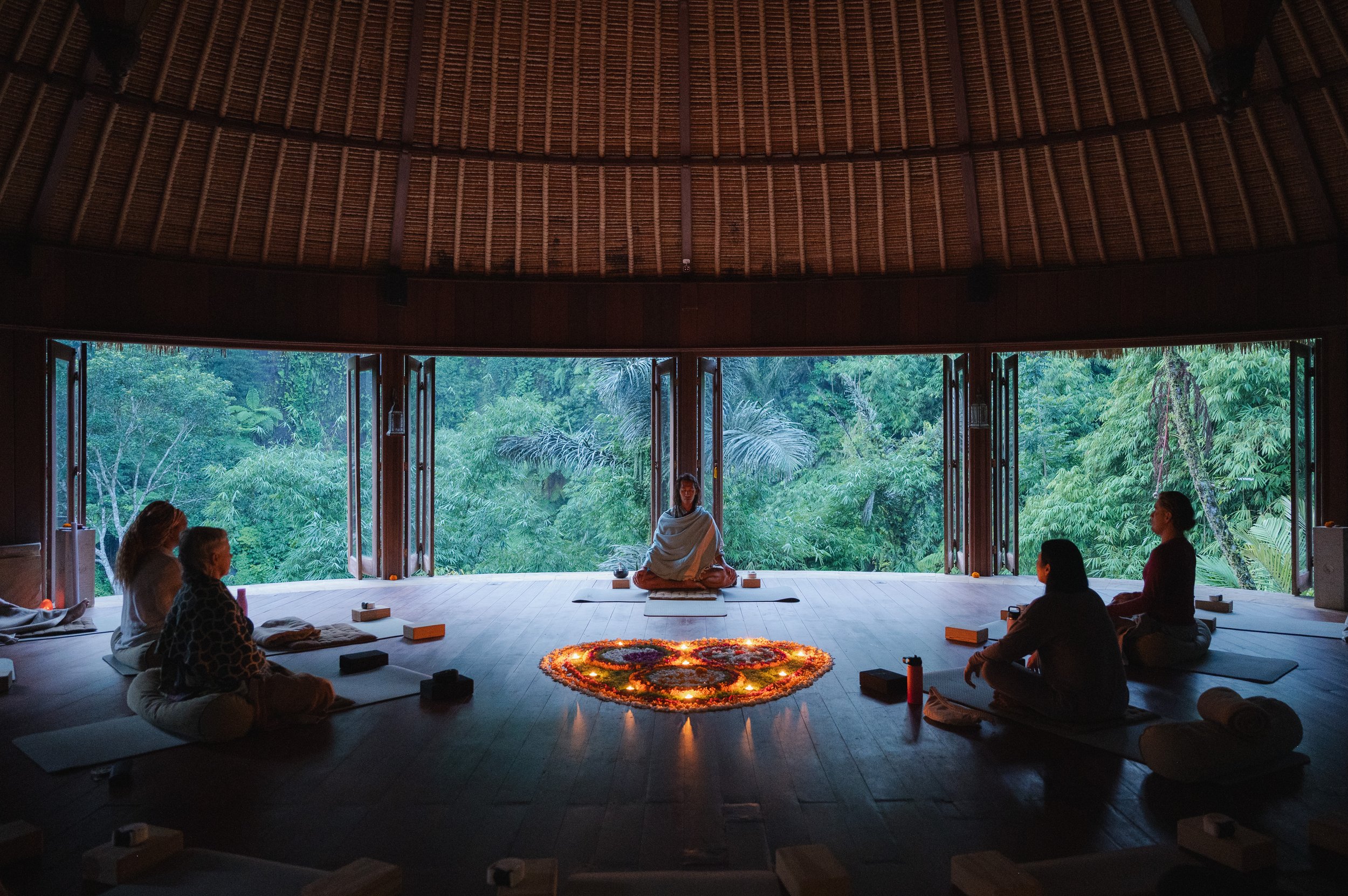Meditation group seated in a tropical retreat with a central instructor, candlelit flower mandala, and lush forest view