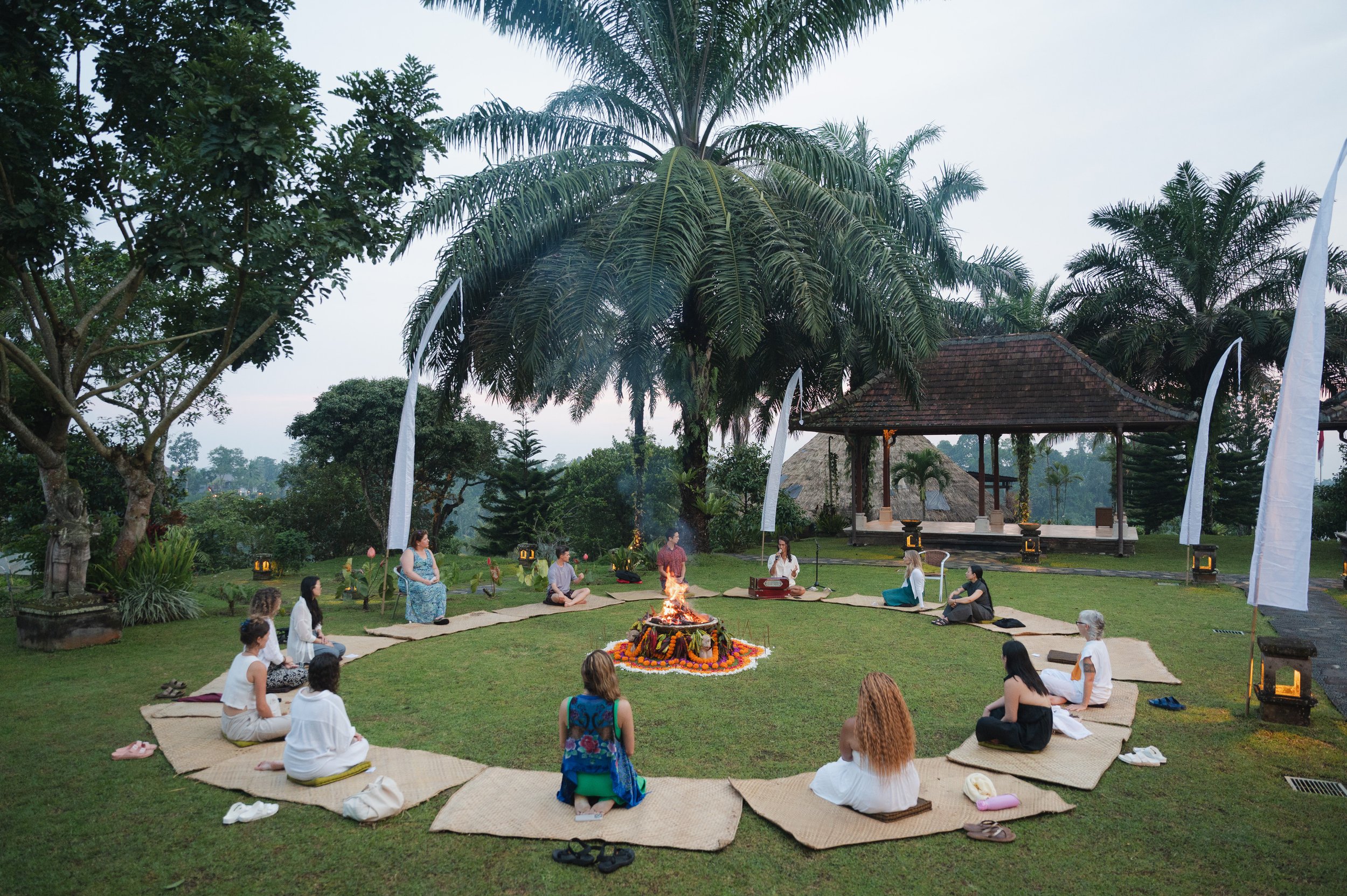 People meditating around a flower-adorned fire altar in a tropical garden with palm trees and a pavilion