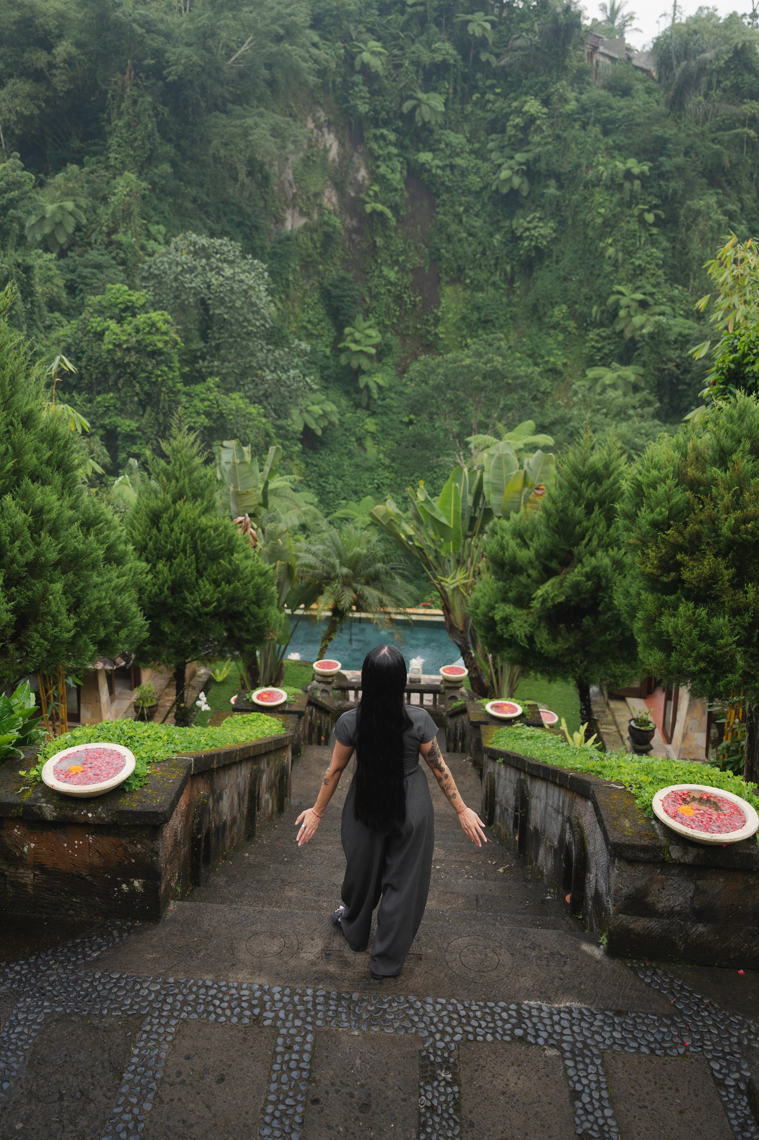 Woman walking down stone steps through a lush tropical garden toward a pool, surrounded by greenery and flower-filled basins.