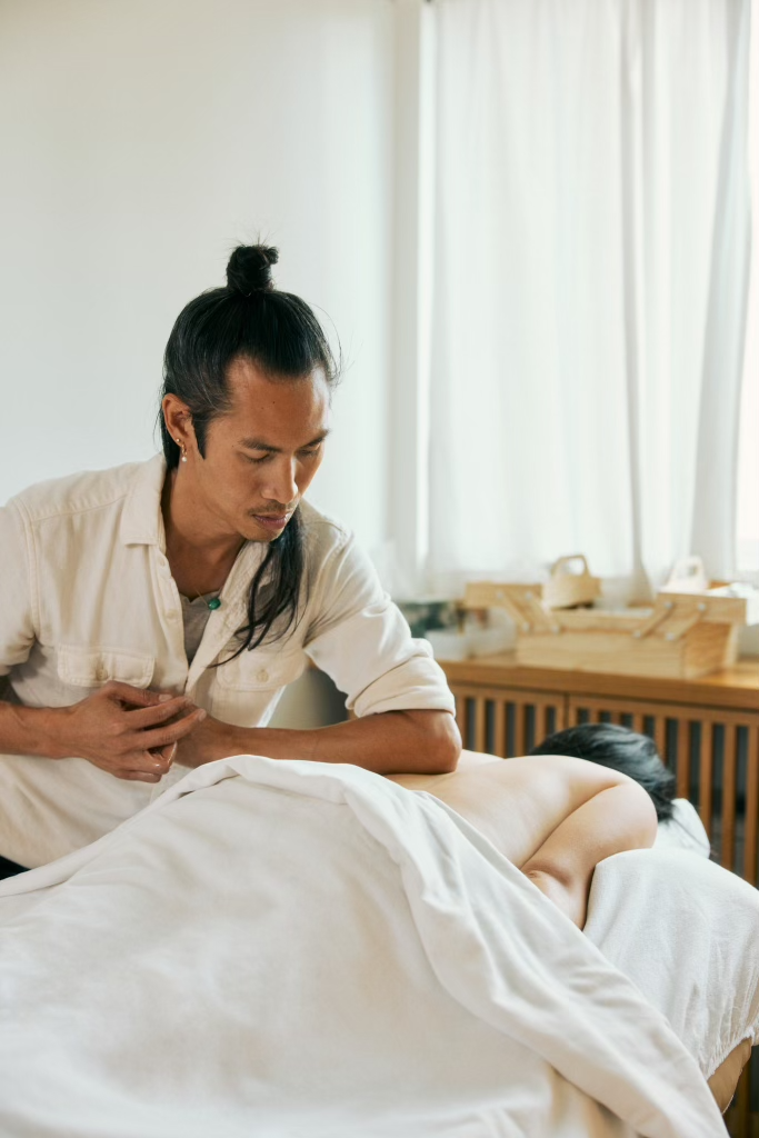 Massage therapist providing a treatment to a client lying under a white sheet in a bright spa room