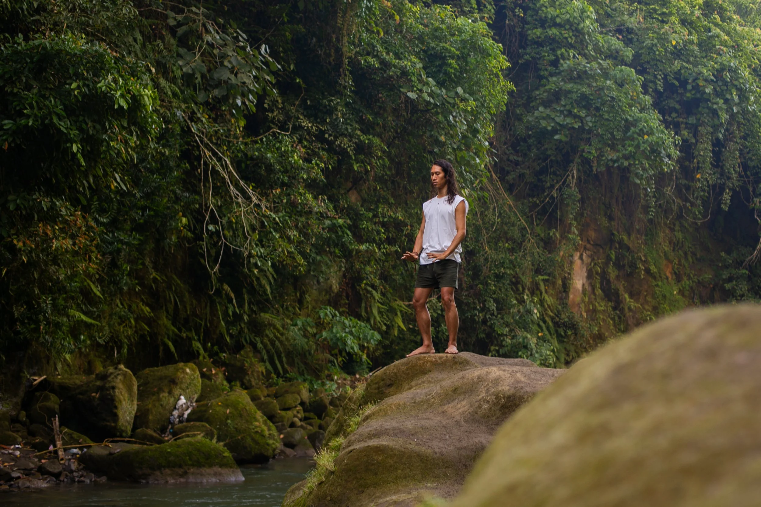 Person practicing meditation barefoot on a rock beside a forest stream