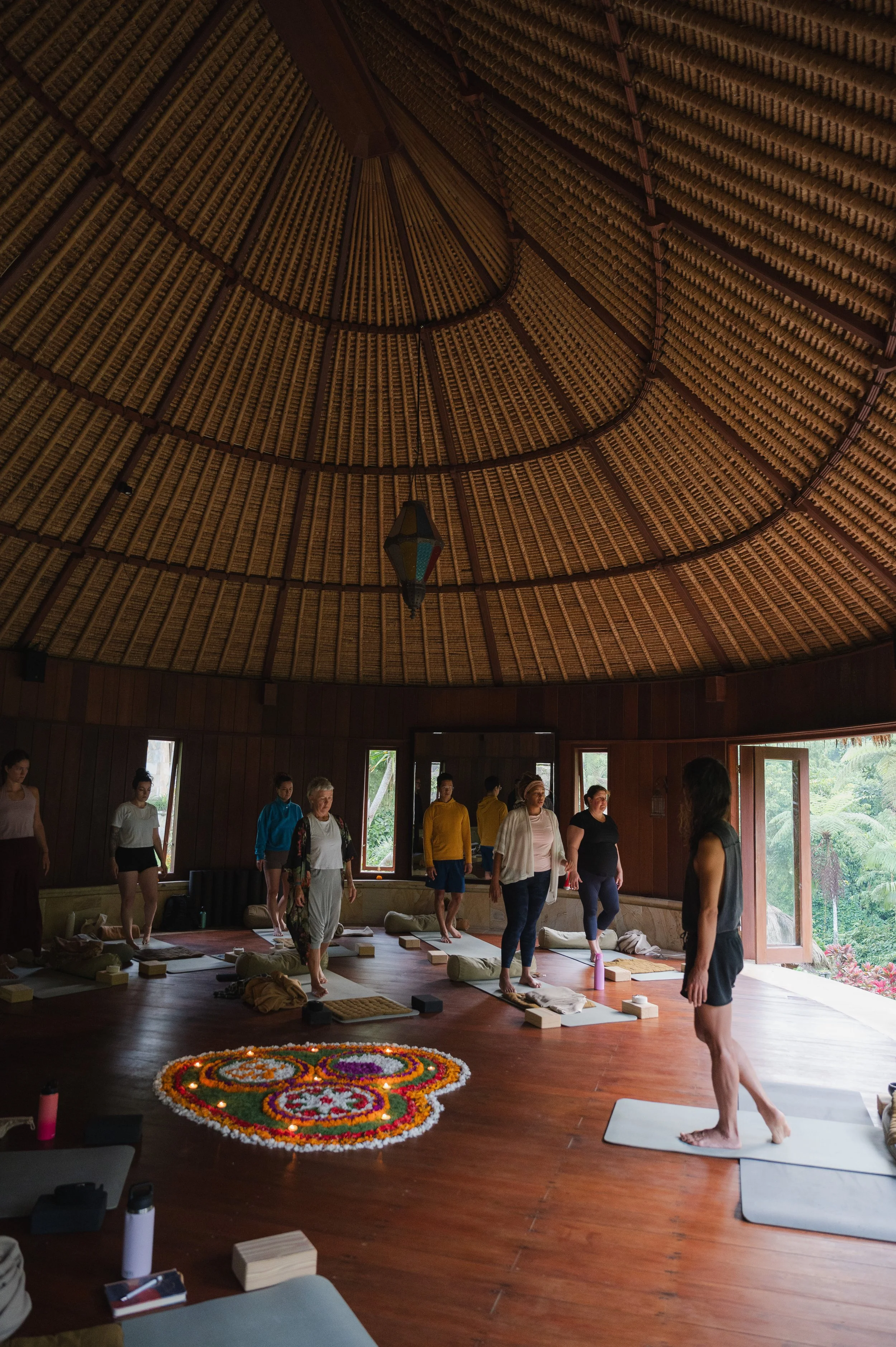 Group yoga class practicing in a rustic meditation hall with a thatched roof, yoga mats, and a colorful flower mandala.