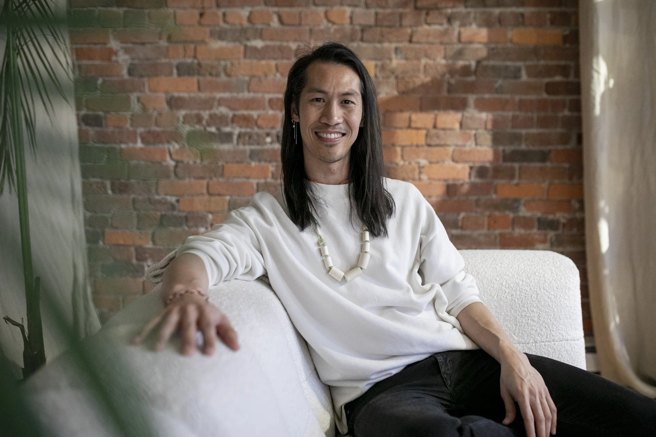 Smiling person with long dark hair seated on a white sofa against a brick wall