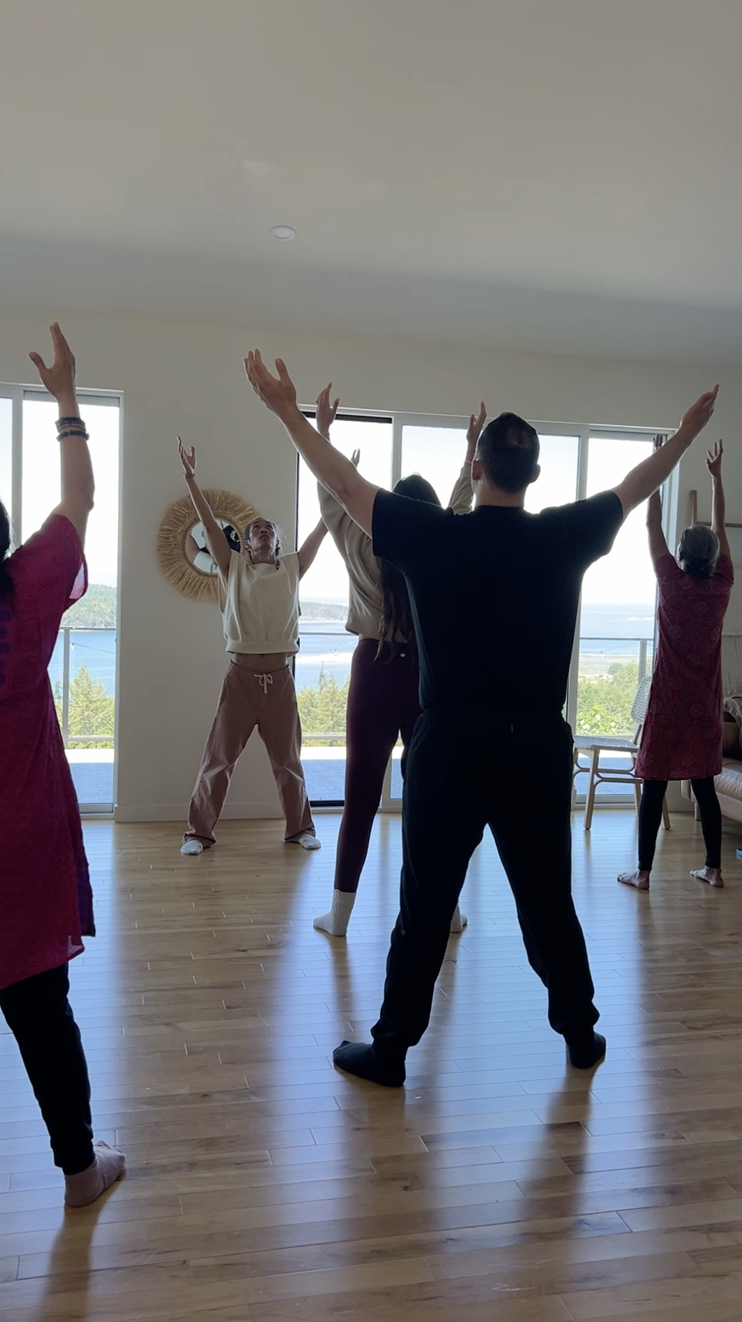 Group of people practicing qigong with raised arms in a bright room overlooking the ocean