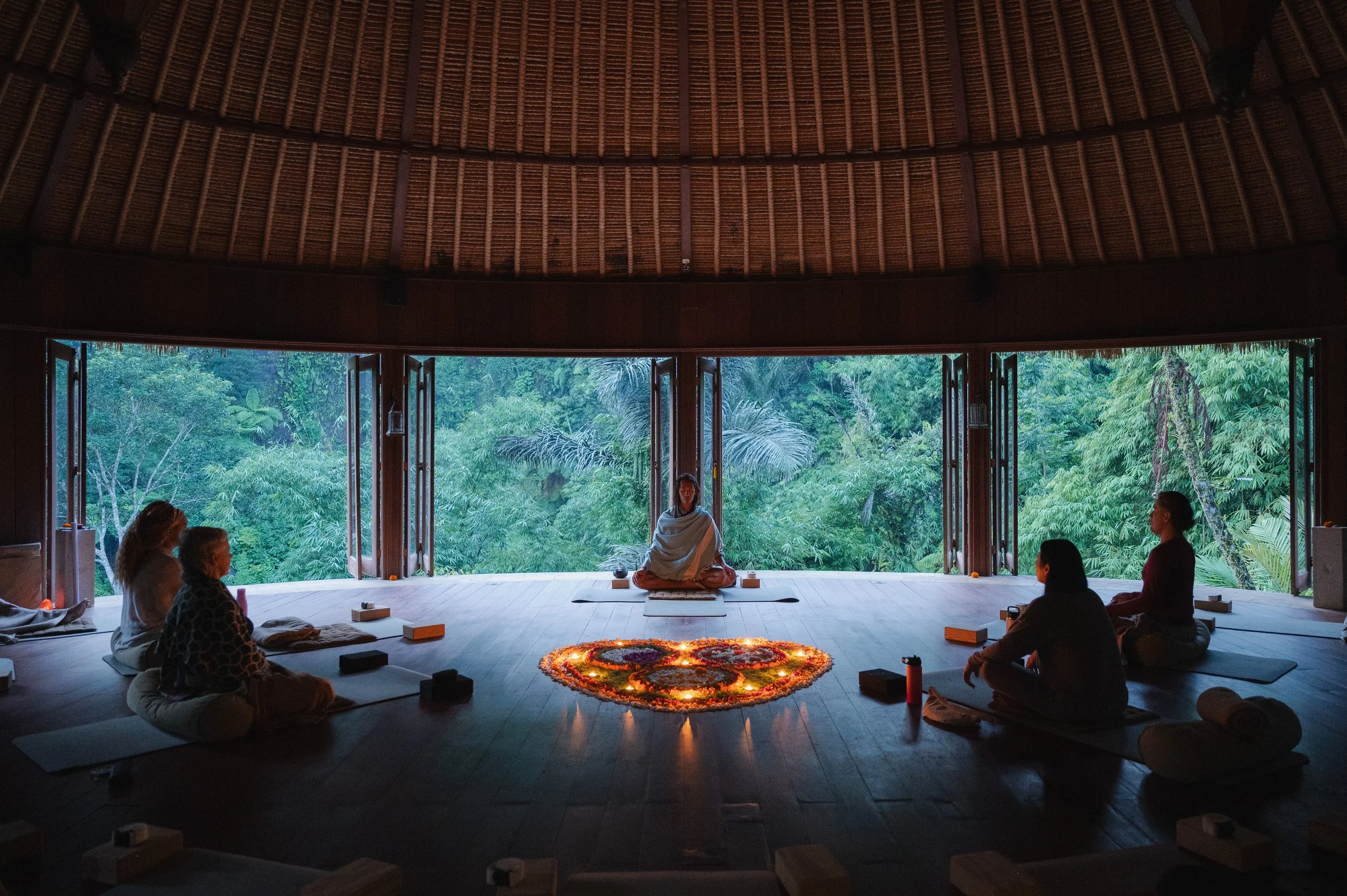 Meditation group seated in a tropical retreat beneath a thatched roof, with a central instructor and illuminated flower mandala.