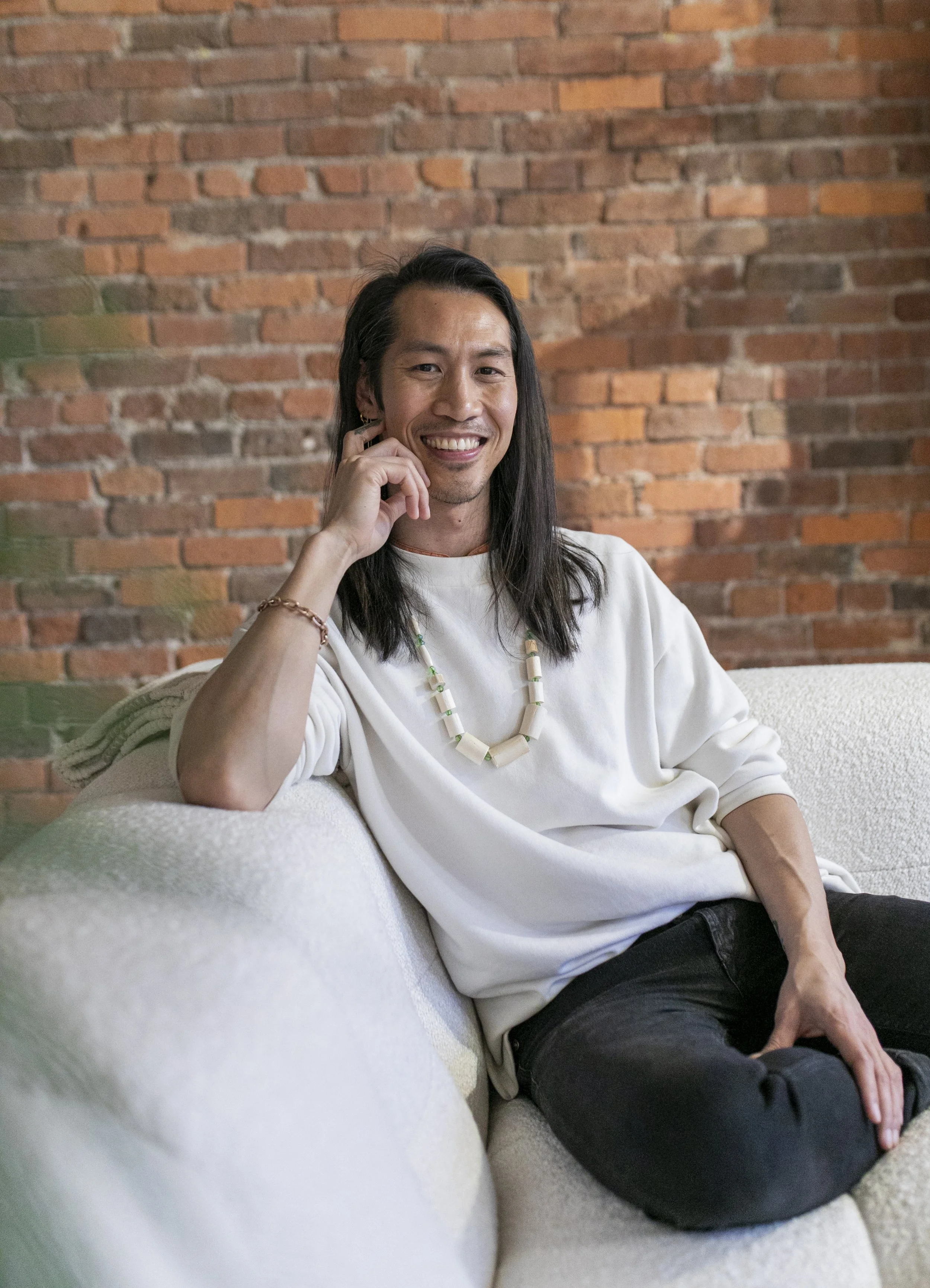 Smiling person with long dark hair wearing a white shirt and beaded necklace, seated on a couch in front of a brick wall