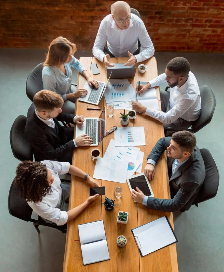 A group of six diverse professionals seated around a conference table engaged in a business meeting, with laptops, documents, phones, and coffee cups on the table.