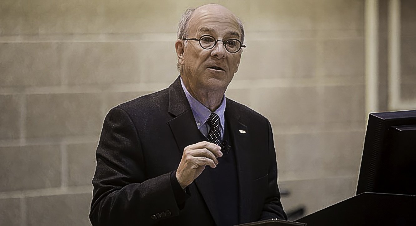 Gray Whale Media Owner Michael Williams, an older man wearing glasses, a dark blazer, a light blue shirt, and a patterned tie, is standing behind a podium, speaking or presenting in front of a beige wall.